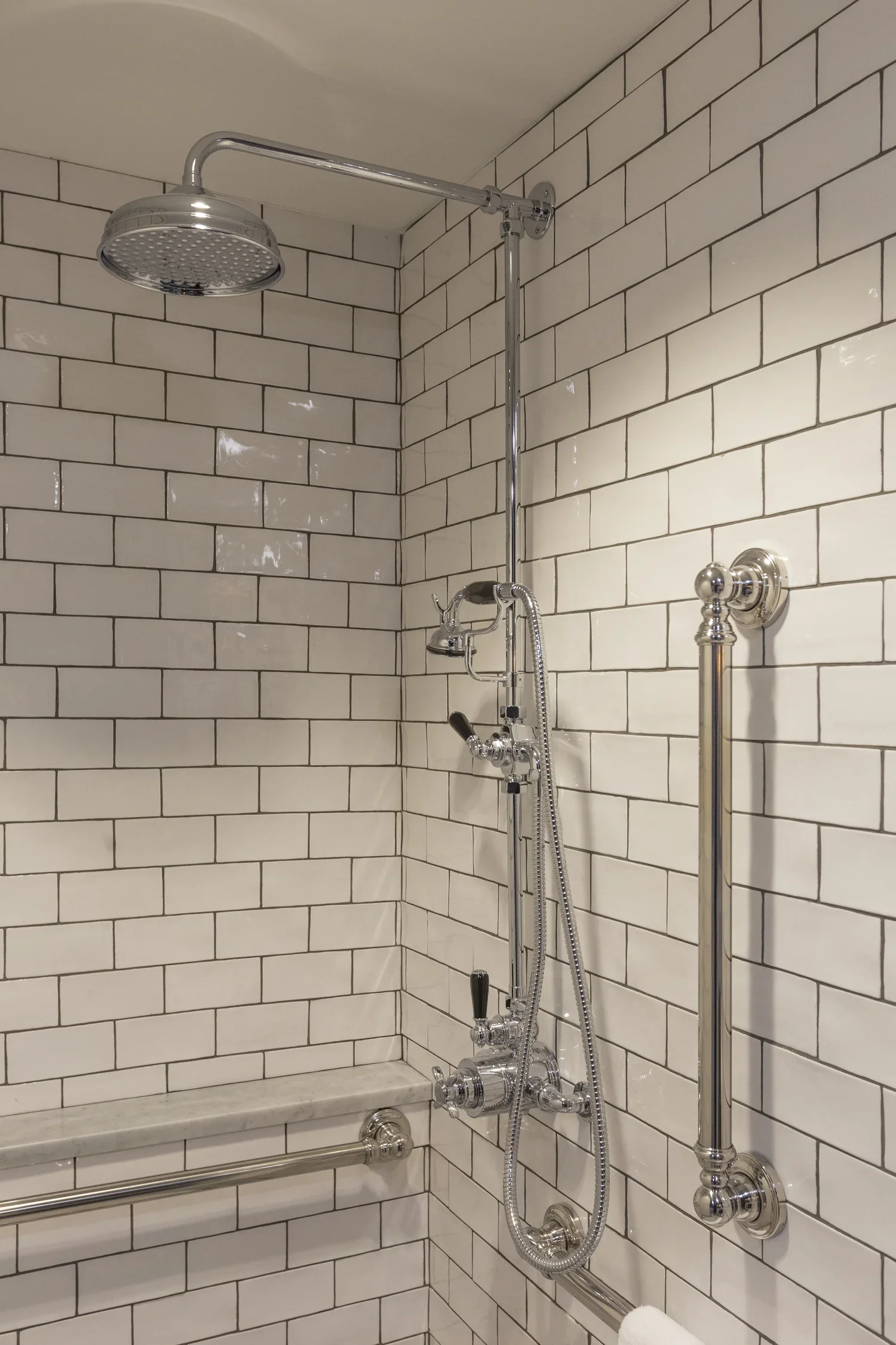 Showerhead and fixtures in a bathroom with white tiled walls and a grab bar.