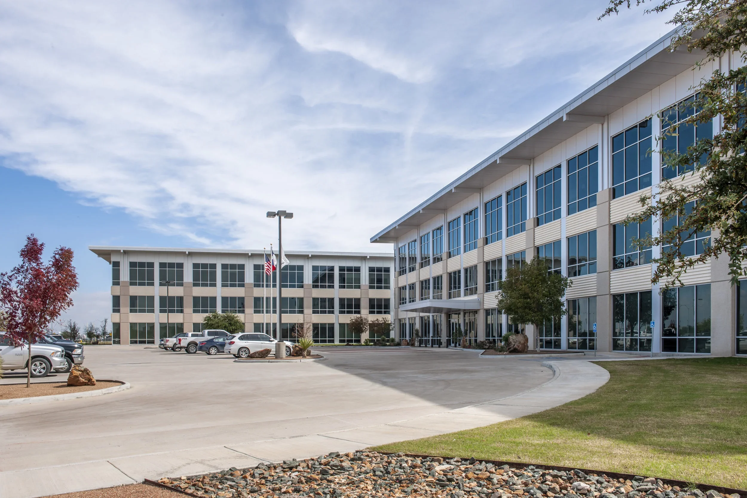 A modern office building with large glass windows, a parking lot with cars, and trees, under a partly cloudy sky.