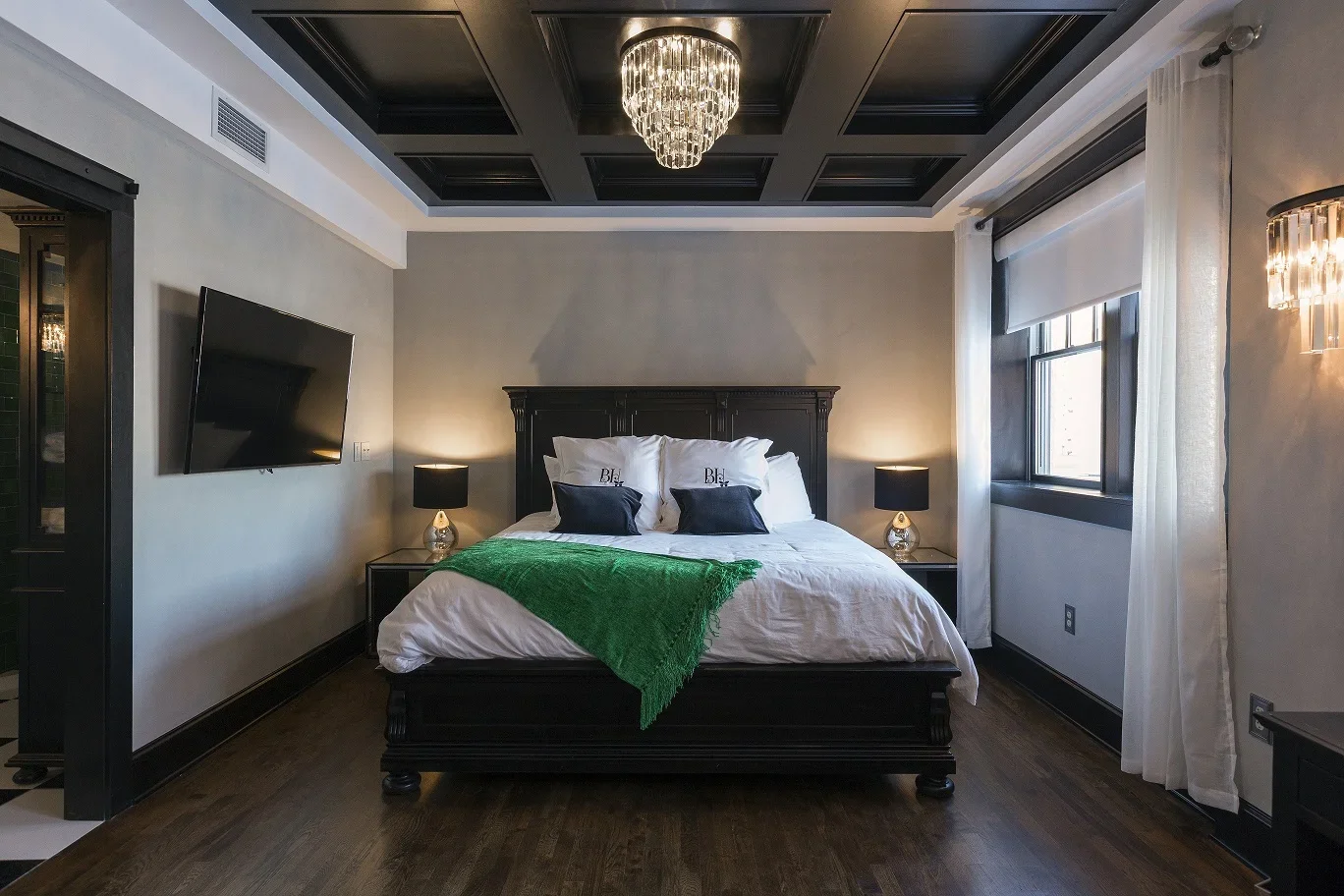 A bedroom with a dark wooden bed, white bedding, black and white pillows, a green blanket, and matching black nightstands with lamps. There's a wall-mounted TV, white curtains, and a window. A chandelier hangs from a coffered ceiling.