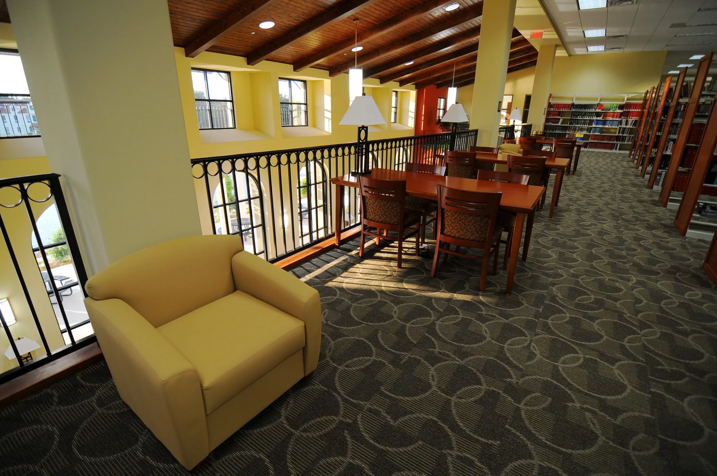 Interior of a library or study area with yellow walls, wooden ceiling, bookshelves, tables, chairs, and a yellow sofa near a black metal railing overlooking a lower level.