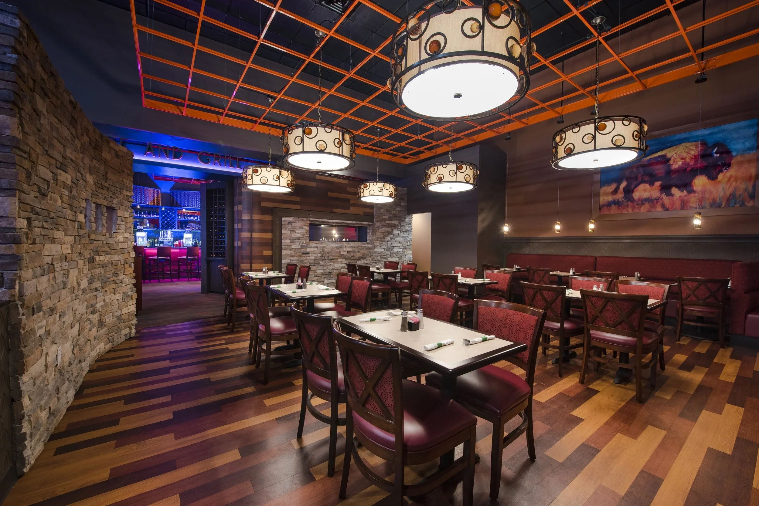 A cozy restaurant interior with wooden flooring, red cushioned chairs, white tables set with napkins and condiments, hanging round lamps, stone and wood accent walls, and a bar area with neon lighting in the background.