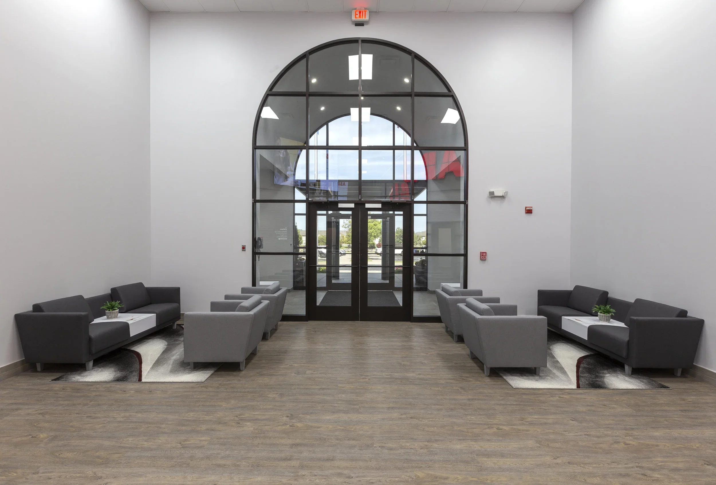 Modern lobby with high white walls, symmetrical gray and black sofas with small potted plants, large arched window and glass doors, wood flooring, fluorescent ceiling lights, visible outside parking lot and trees.