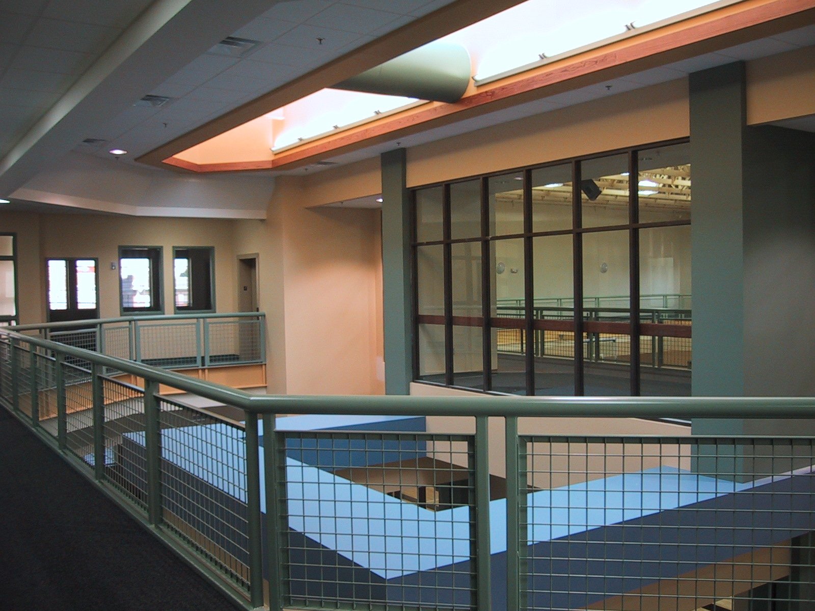 Indoor hallway with teal railings and large glass window looking into a fitness room or gym with exercise equipment.
