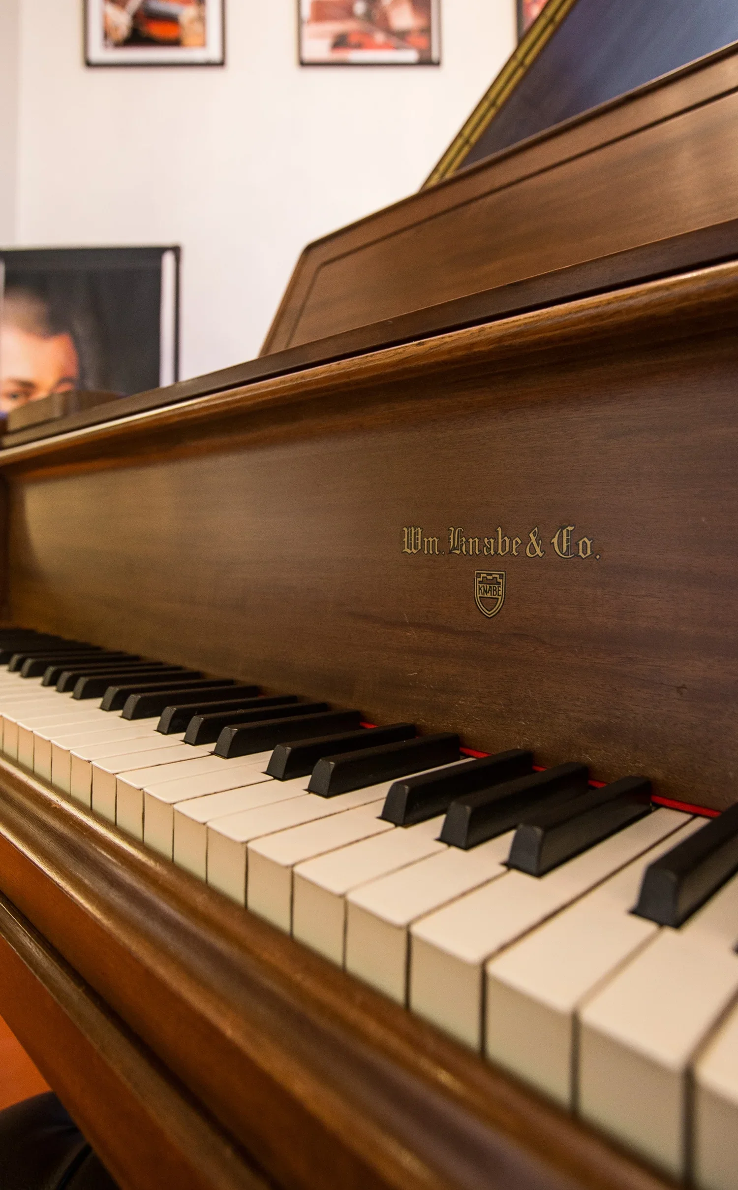 Close-up of a wooden upright piano with black and white keys in a room with framed pictures on the wall.