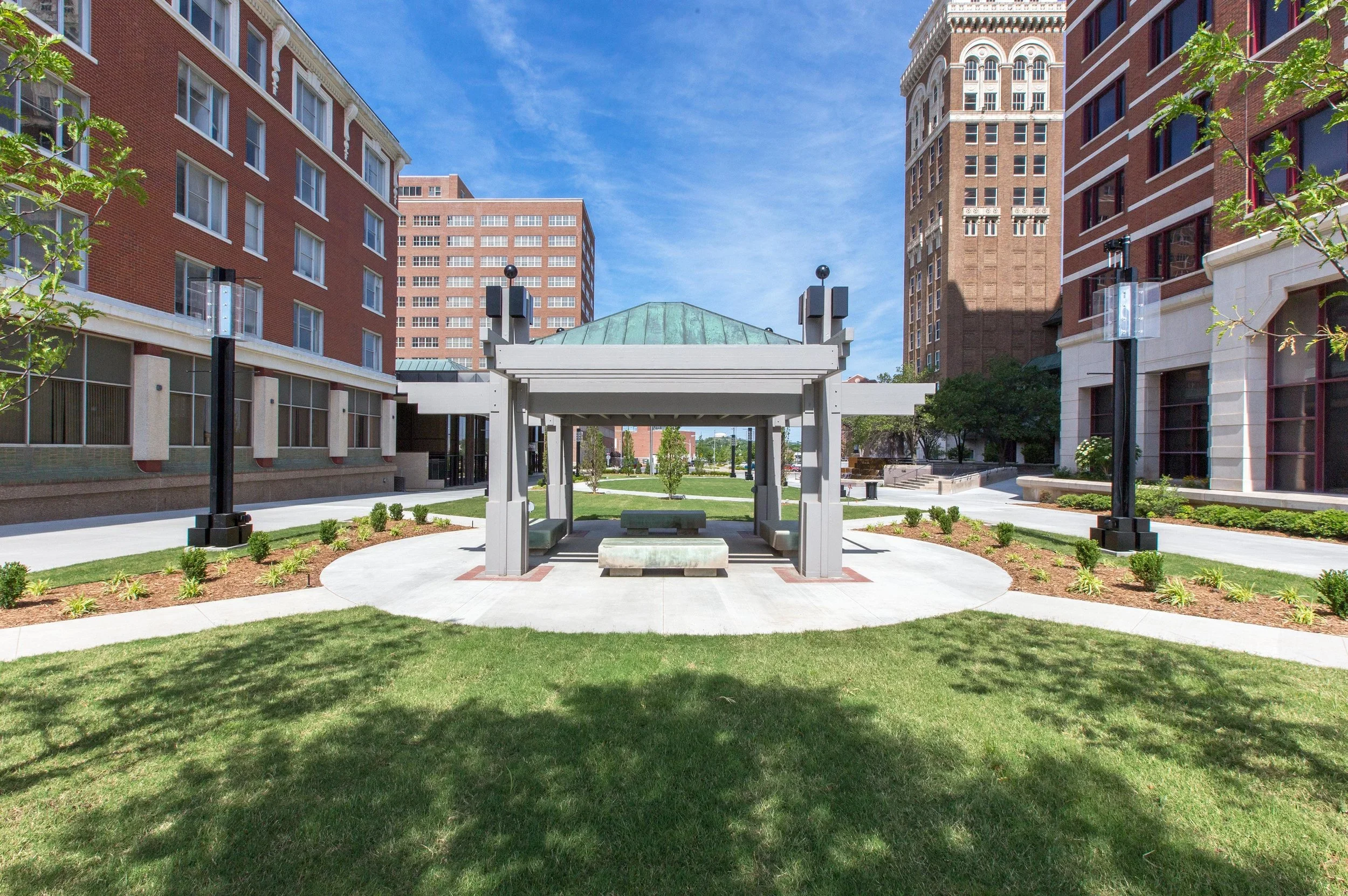 A small pavilion with a green roof in the center of a landscaped urban park, surrounded by modern brick buildings, with a blue sky overhead.