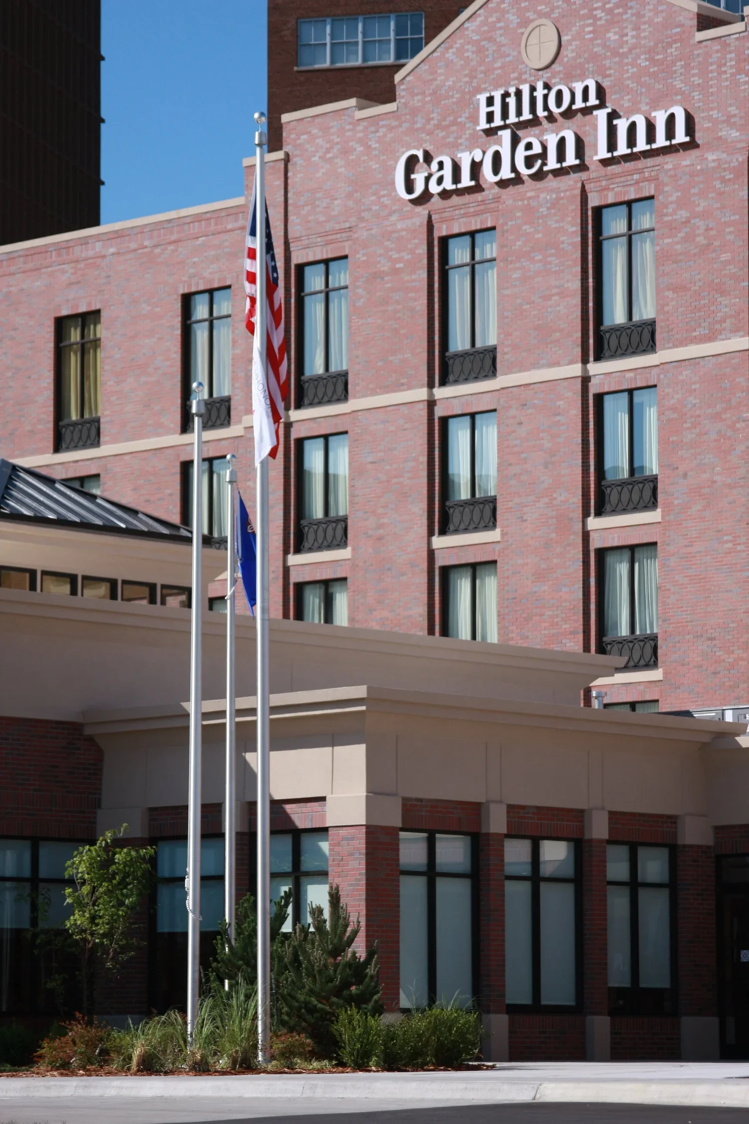 Exterior view of Hilton Garden Inn hotel with flagpoles and flags in front, brick building with multiple floors, windows, and hotel sign at the top.