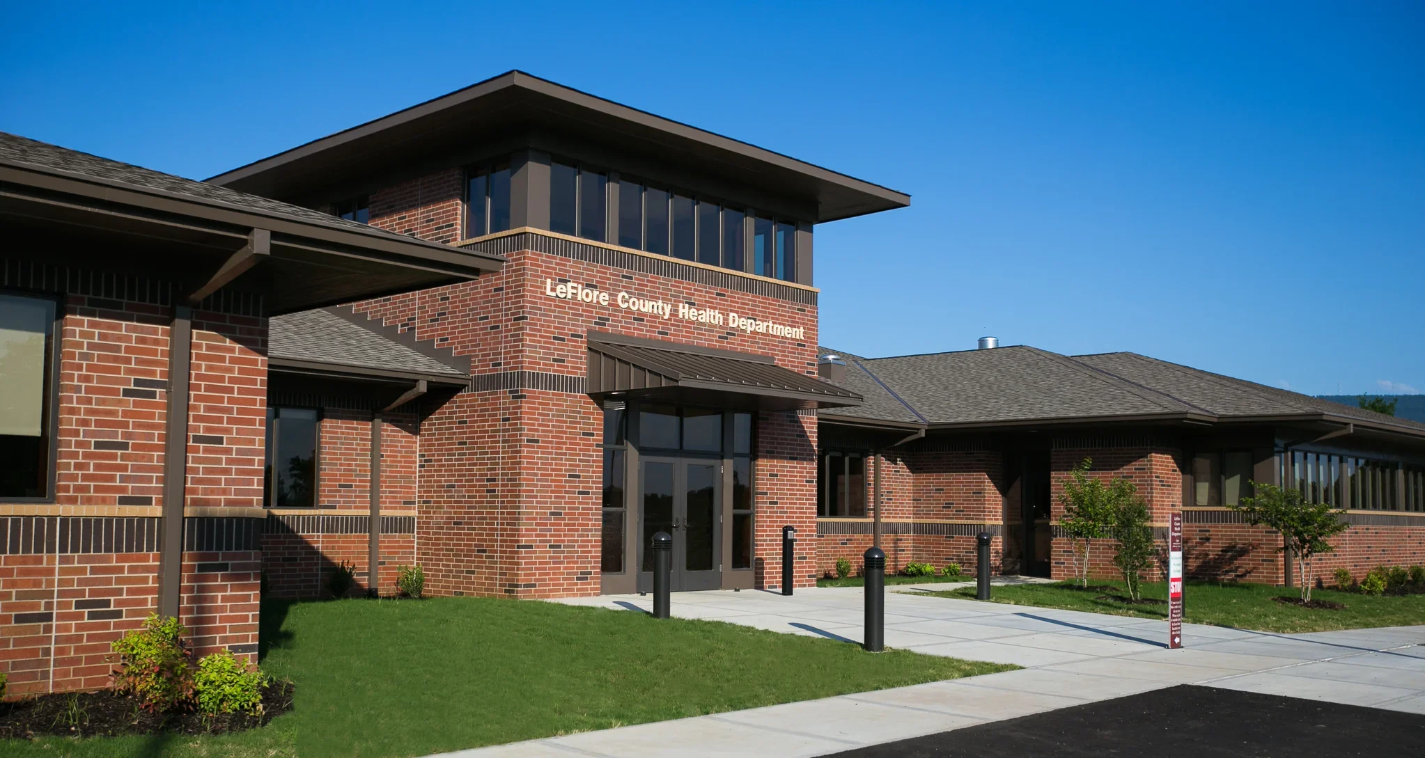 Modern brick building with a sign reading LeFlore County Health Department, surrounded by a lawn and small trees, under a clear blue sky.