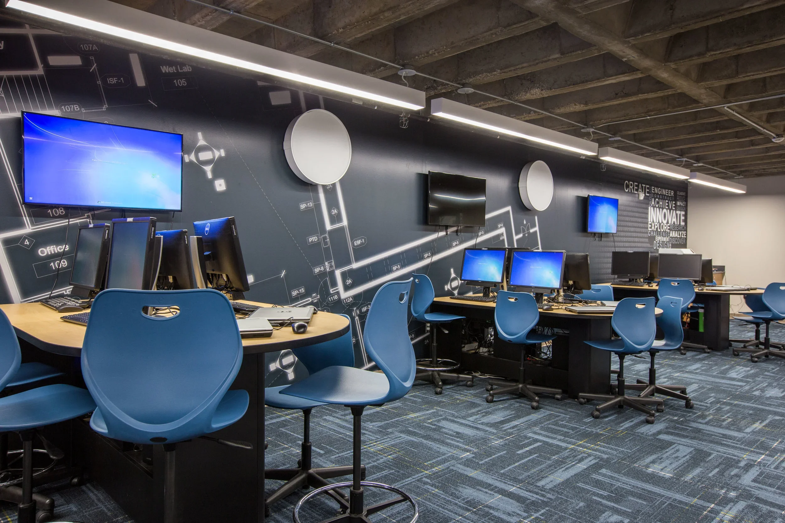 Technology-focused classroom or training room with multiple desktop computers and monitors, blue chairs, and a modern wall with diagrams and motivational words.