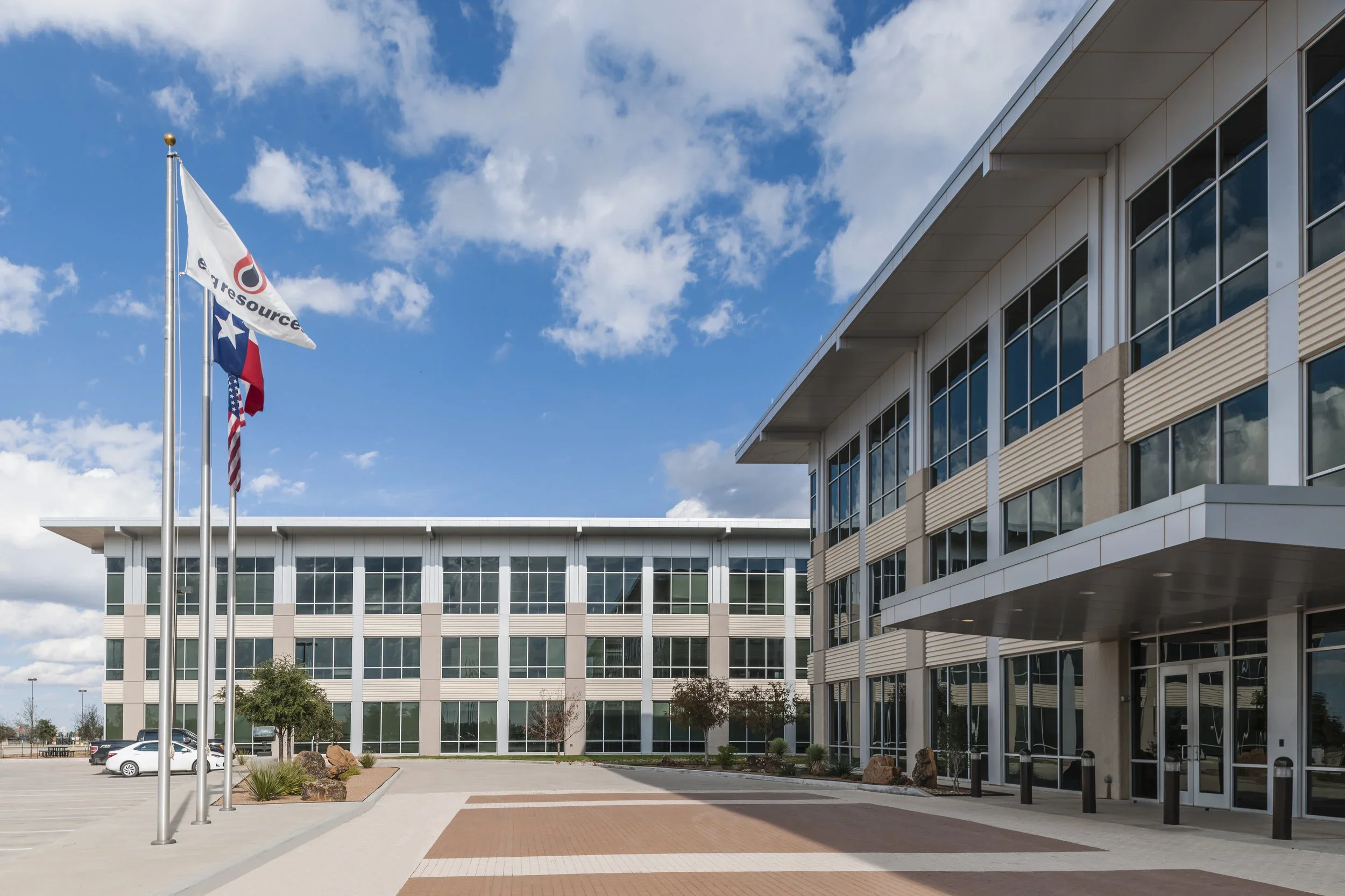 Modern office building with large glass windows, flagpoles with flags, and a parking lot on a sunny day.