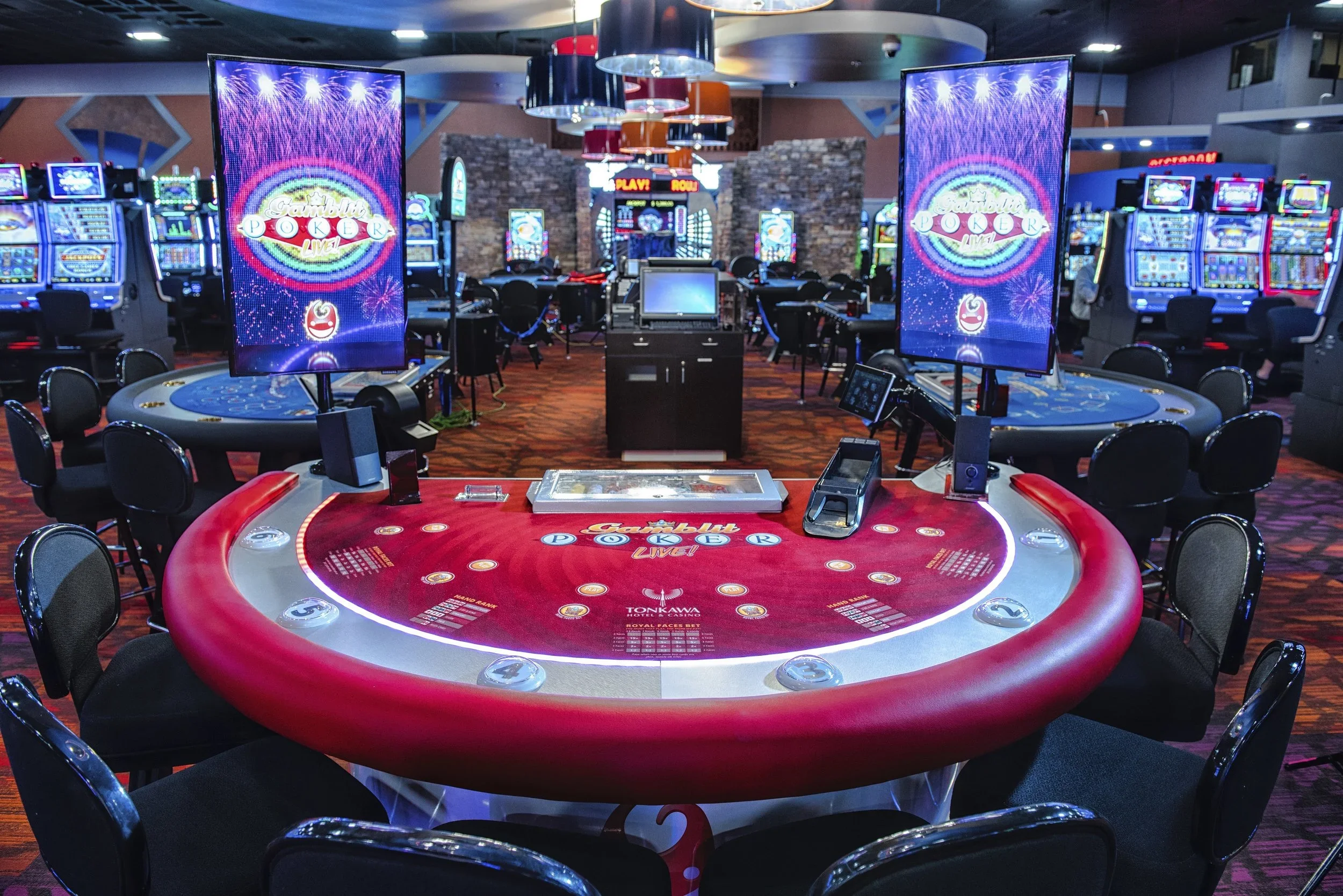 Inside a casino with gaming tables, electronic gambling machines, and large screens displaying colorful graphics and fireworks.