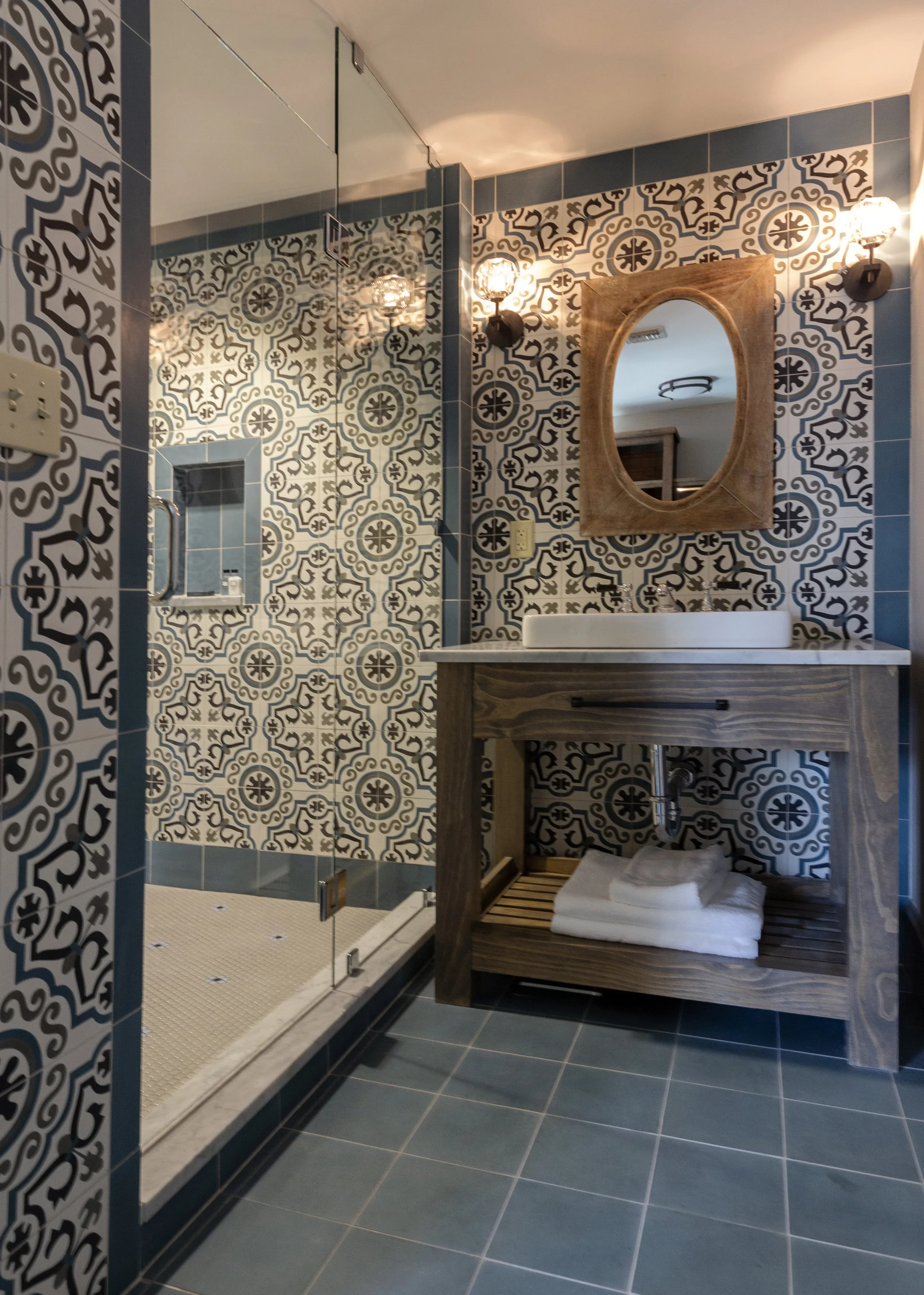 A bathroom with patterned black and white tiles on the wall, blue floor tiles, a wooden vanity with a vessel sink, a mirror with a wooden frame, and a glass-enclosed shower with a niche and a small shelf.