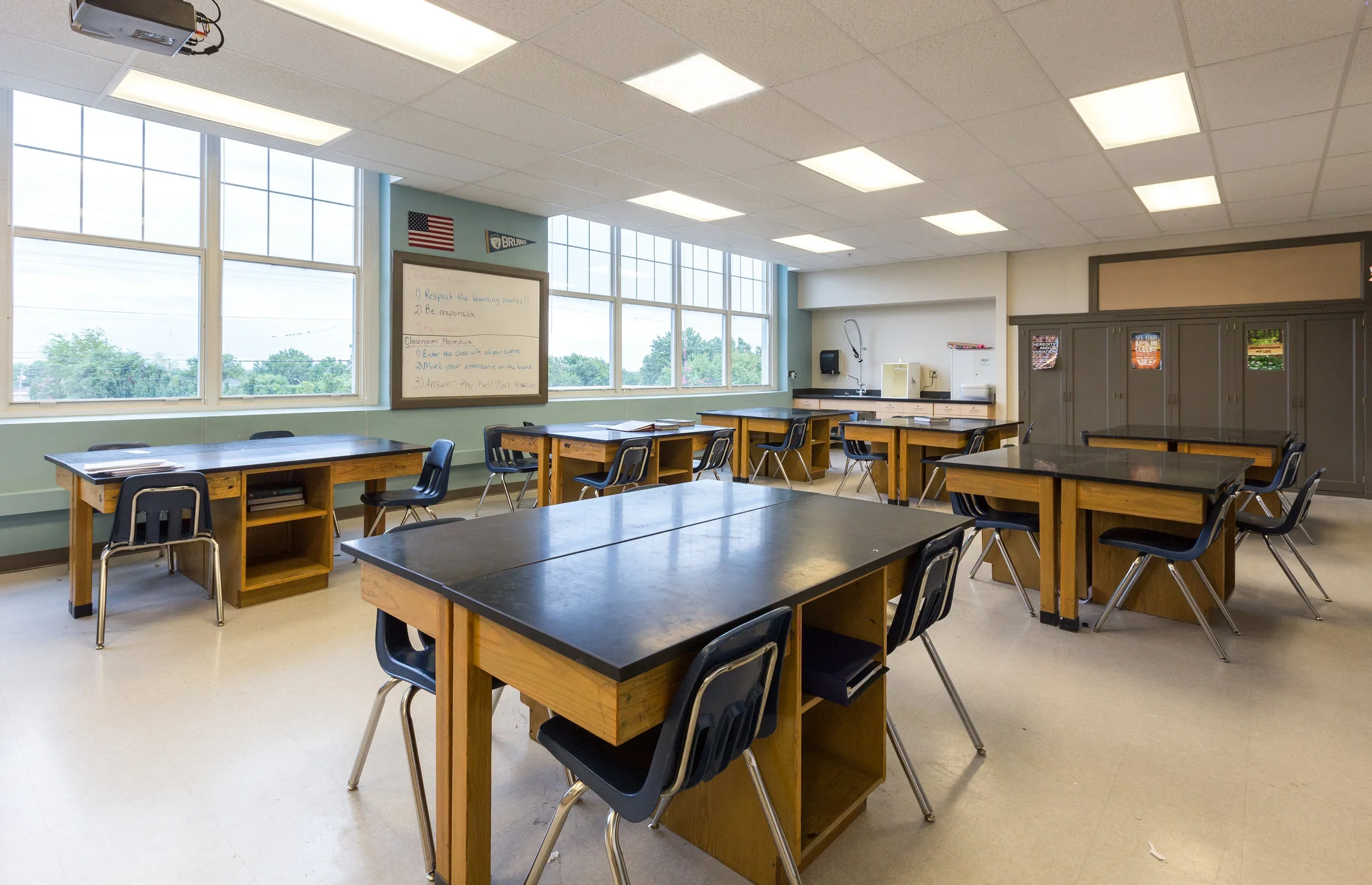 Empty classroom with wooden tables, black chairs, large windows, a whiteboard, and storage cabinets.