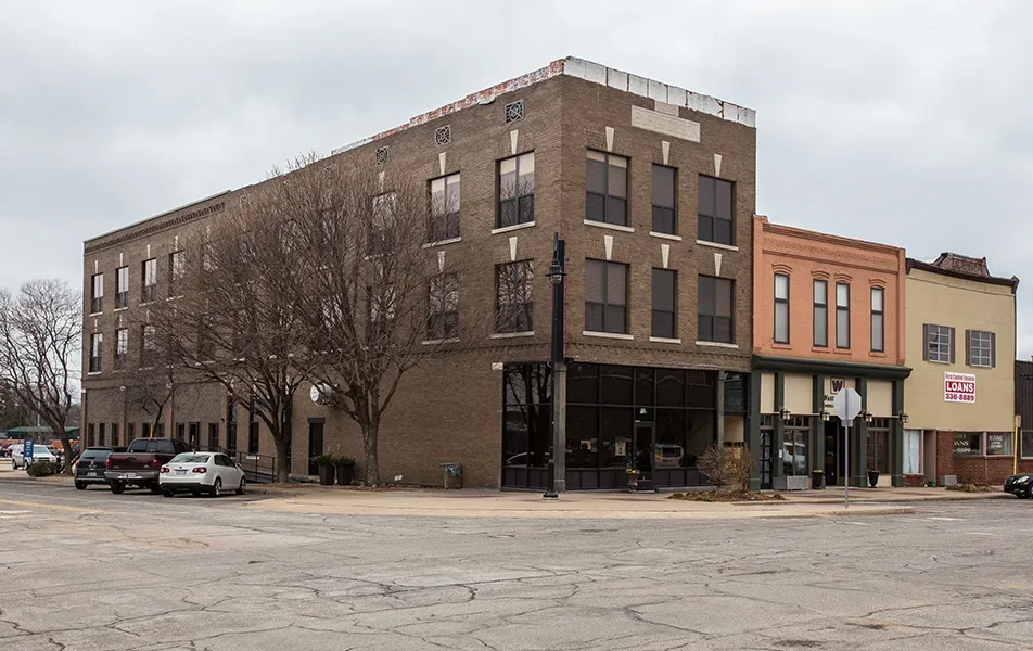 A three-story brick building on a corner with parked cars in front, leafless trees, and a neighboring two-story building with a sign advertising loans.