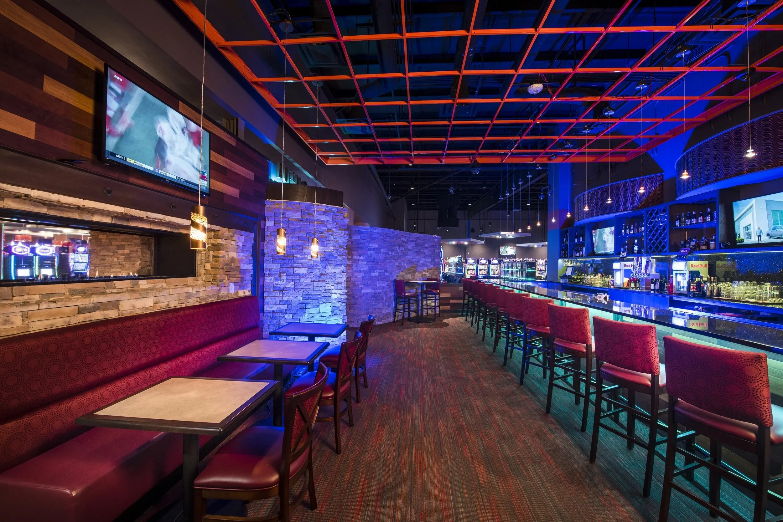 Empty bar with red cushioned chairs, tables, and a long bar counter with illuminated shelves of bottles, neon signs, and multiple flat screens displaying sports.