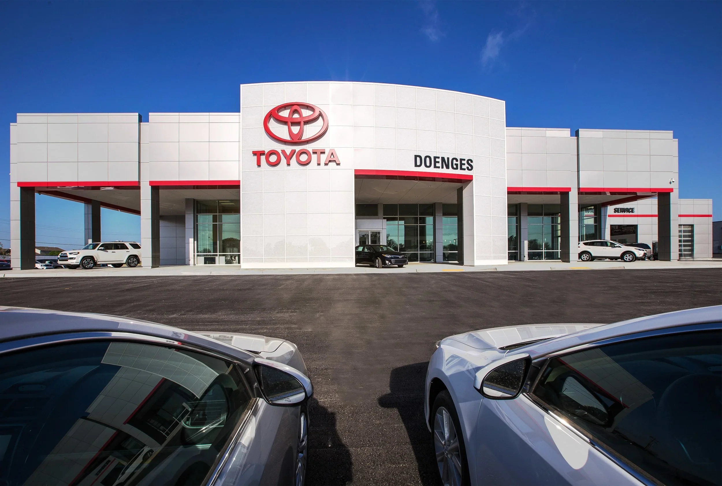 Exterior view of a Toyota dealership building with a large Toyota logo and signage, parked cars in front, under a clear blue sky.