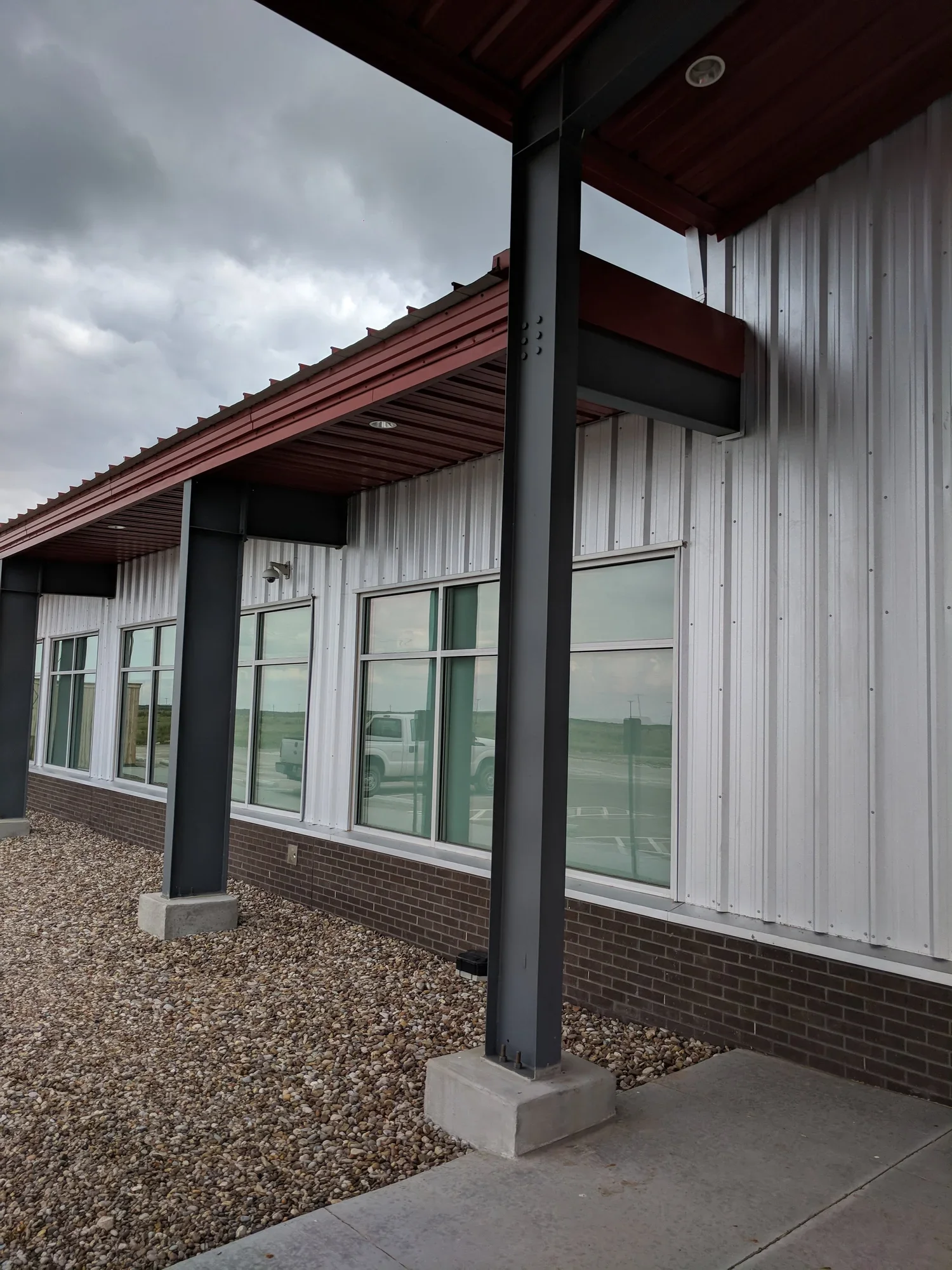 Exterior view of a building with metal siding, large windows, and steel support columns on a gravel surface under a cloudy sky.