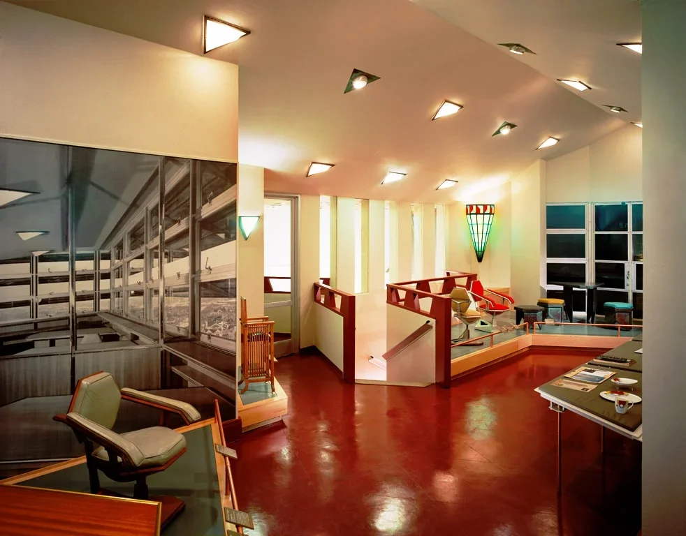 Interior of a modern building with patterned red floor, white walls, ceiling lights, art deco wall sconce, large windows, and seating area with chairs and tables.