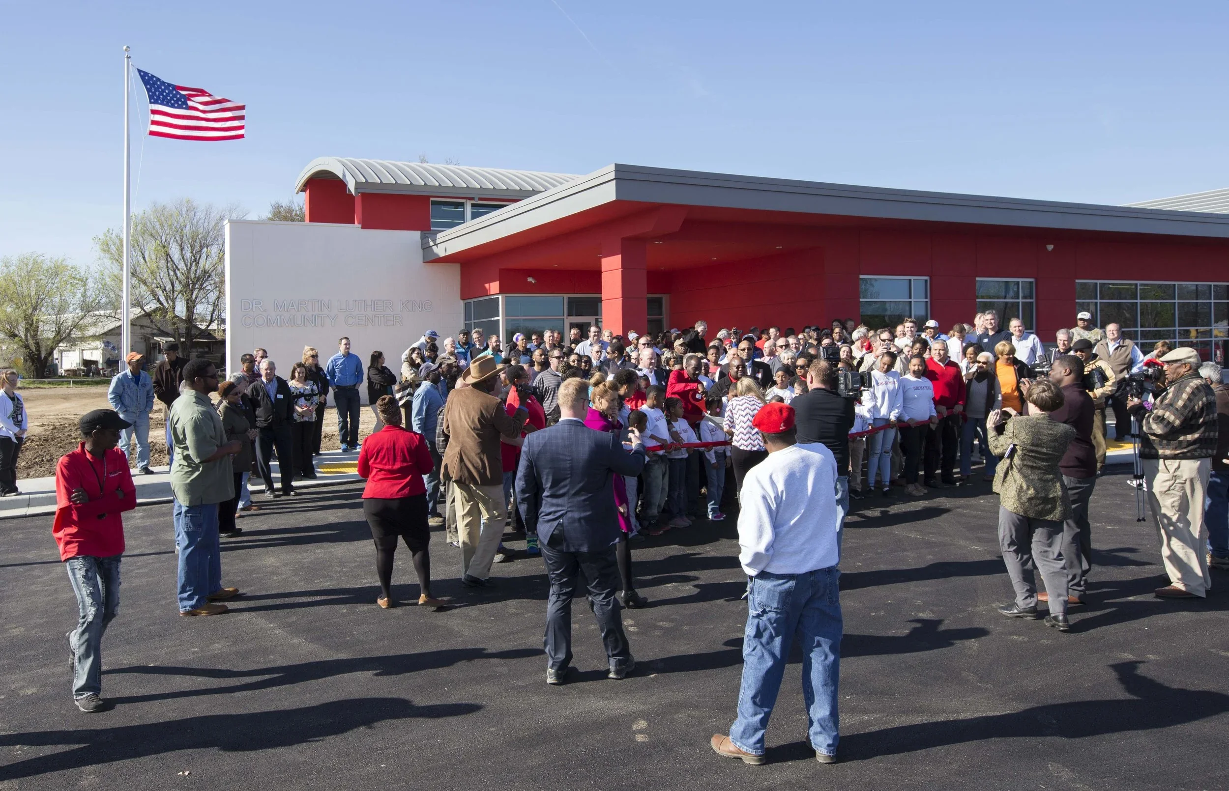 A crowd of people gathered outside the Dr. Martin Luther King Community Center, with some speaking or observing during a ceremony, under an American flag on a bright day.