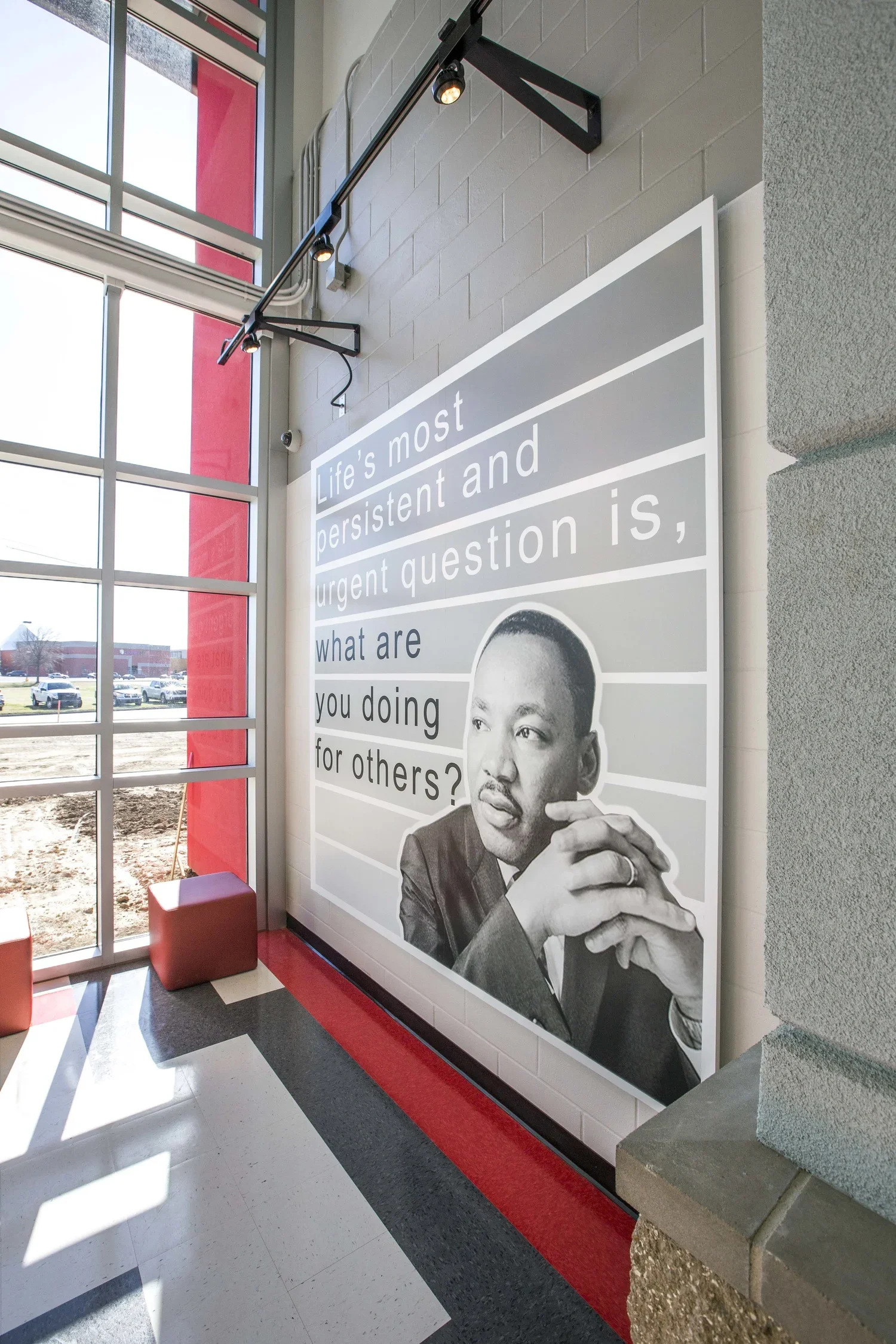 A large wall poster with a black and white photo of a man in a suit and a quote that reads, 'Life's most persistent and urgent question is, what are you doing for others?' inside a modern building with large windows, red accents, and seating.