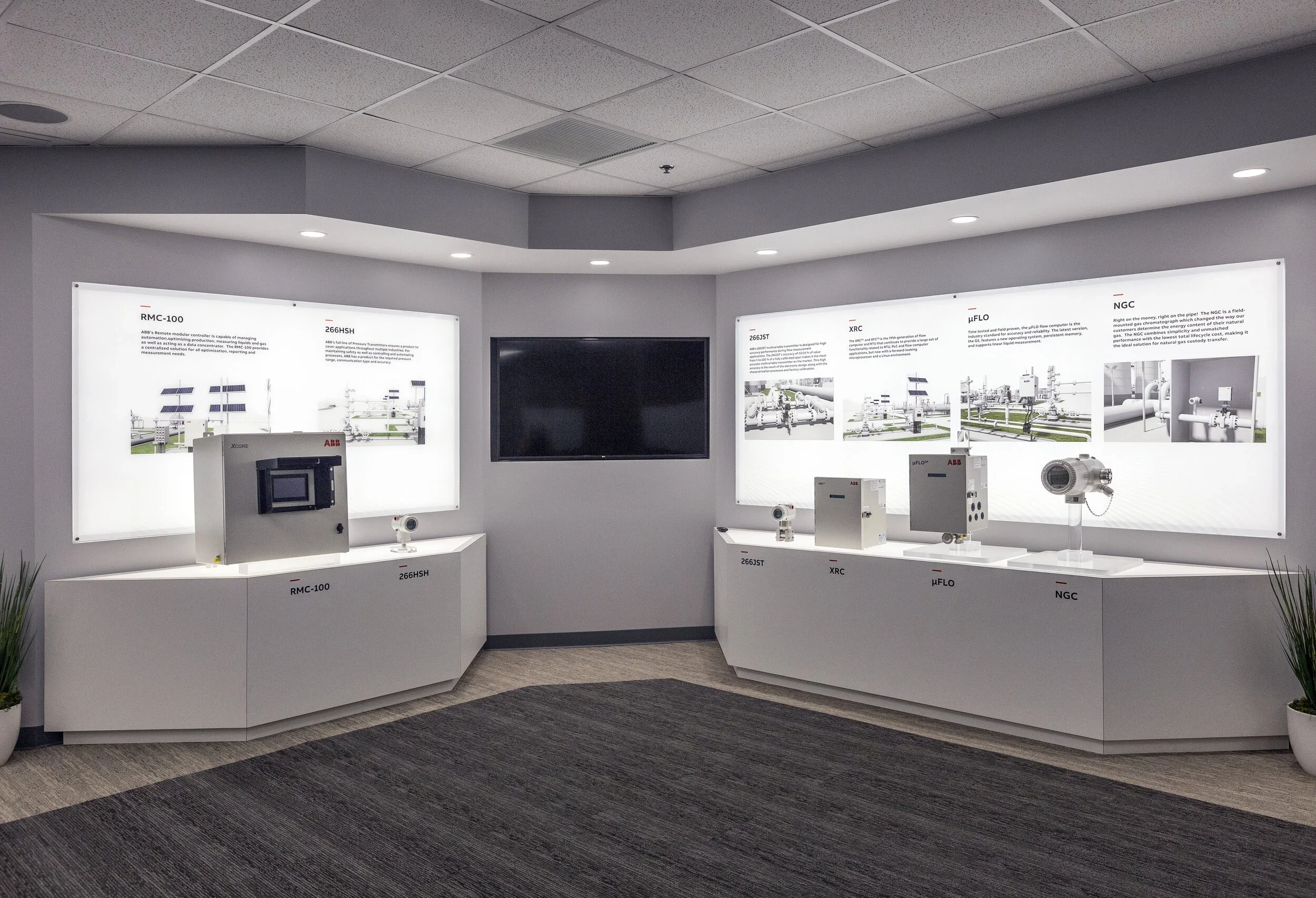 An exhibition display of industrial equipment and informational posters with diagrams and descriptions. Four devices are placed on white display cabinets in front of illuminated panels with technical information and illustrations in a corporate setti