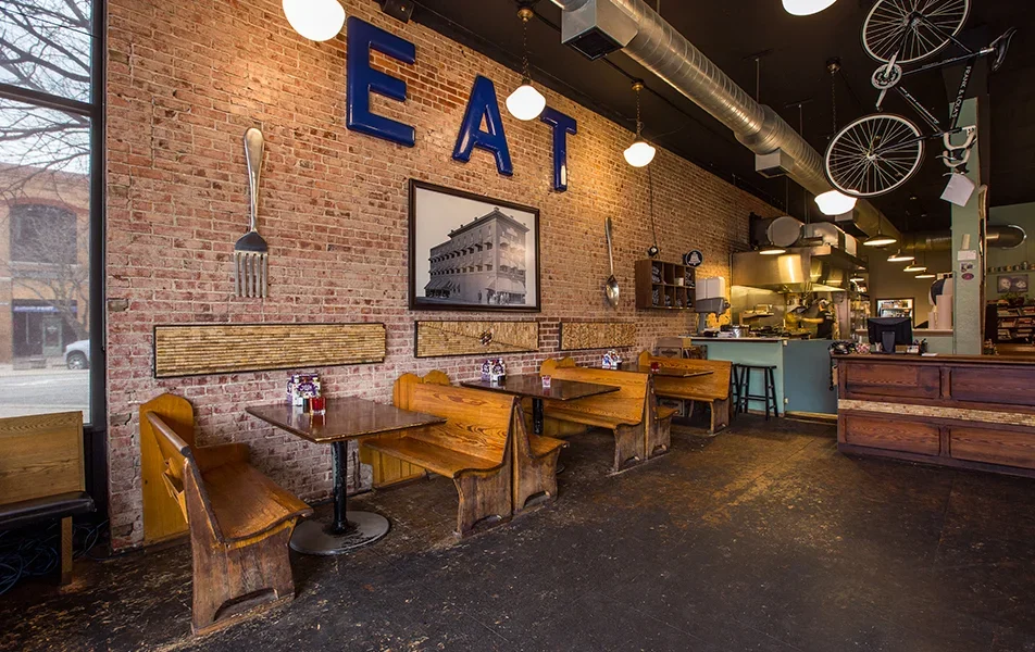 Interior of a restaurant with wooden tables and benches, exposed brick wall with large blue letters spelling 'EAT,' and decorative utensils. The cafe has large windows, industrial-style ceiling with ductwork, hanging lights, and a partial view of the