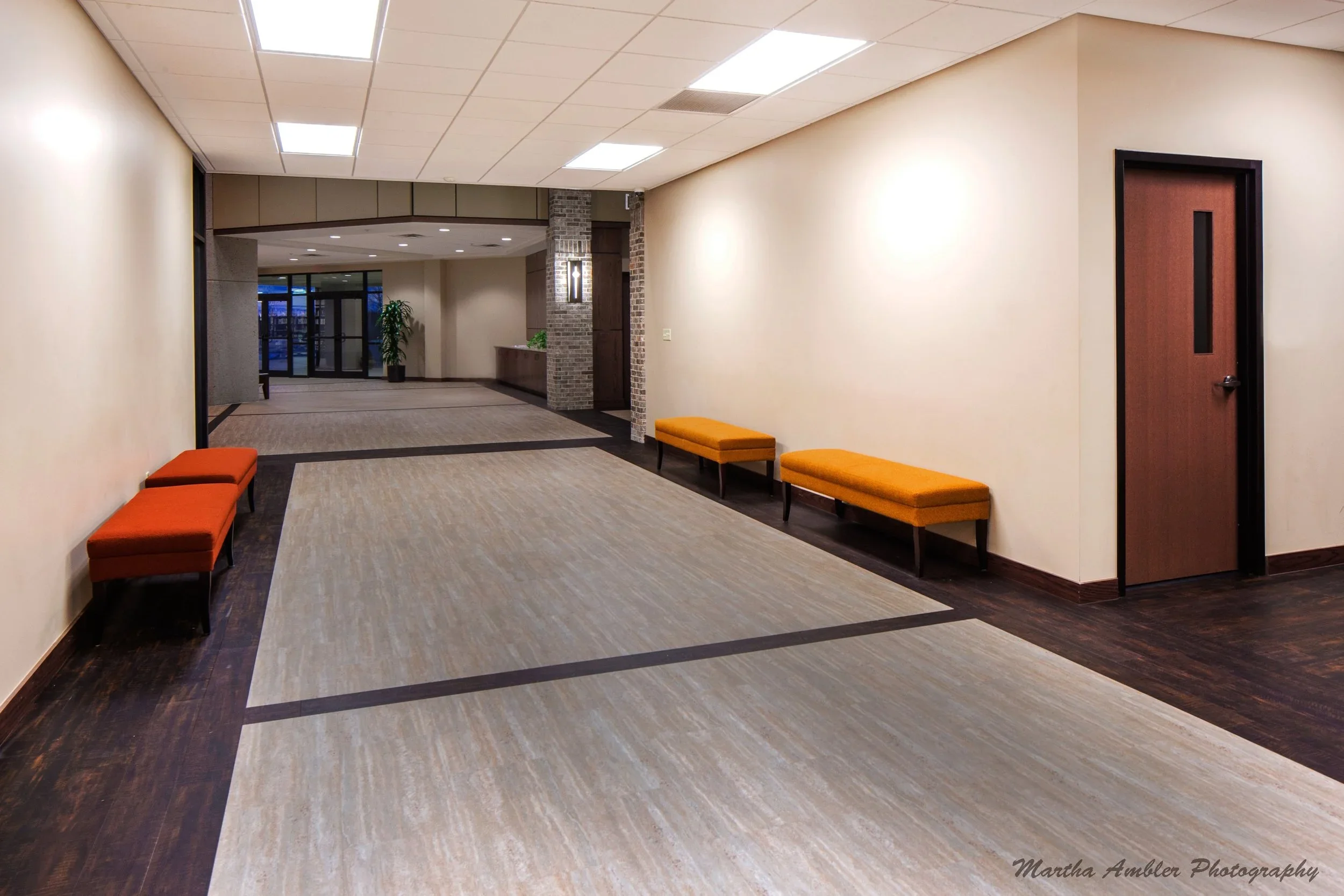 Empty hallway in a building with beige walls, wooden doors, and benches with orange cushions along the wall. The floor is a mix of dark wood and light-colored carpet. There is a plant near the glass entrance doors in the background.