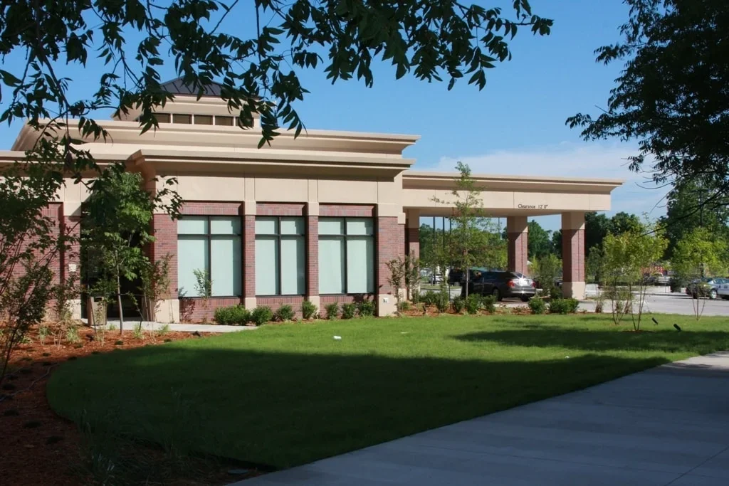 A modern commercial building with large windows, surrounded by landscaping, trees, and a parking lot with cars.