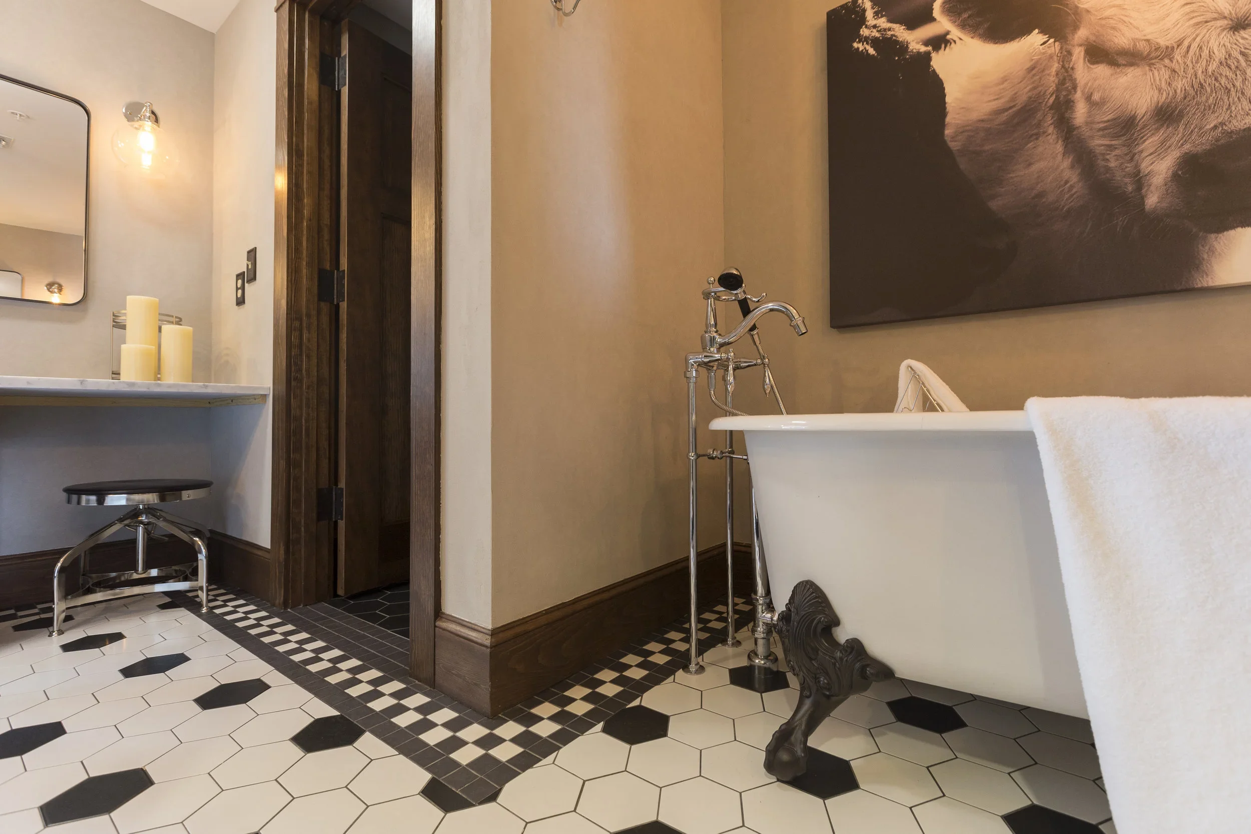 A vintage clawfoot bathtub with a white towel draped over its side, standing on black clawfoot legs, with a chrome faucet, in a bathroom with black and white hexagonal tile flooring and beige walls, a large black and white animal portrait, and a part