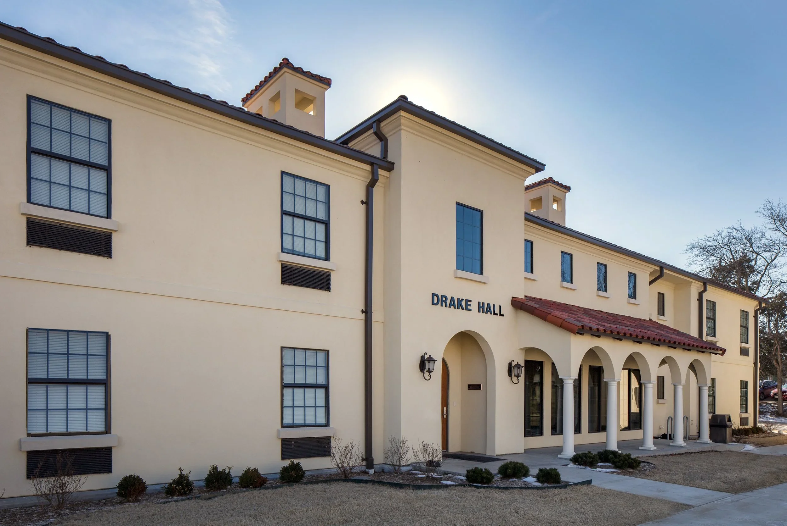 Exterior of Drake Hall building with cream-colored walls, black window frames, and a red-tiled roof, featuring an arched entrance with columns and a sign that reads 'DRAKE HALL' under a clear sky.