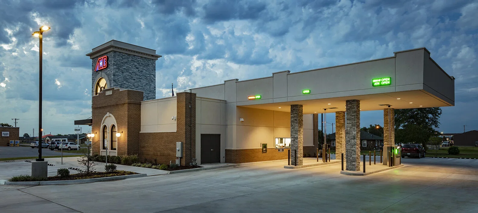 A drive-thru bank building at dusk with illuminated signs saying 'Open' and 'ATM Open', brick and stone design, a tall clock tower with the ABC logo, street lamps, parking lot, and parked cars.
