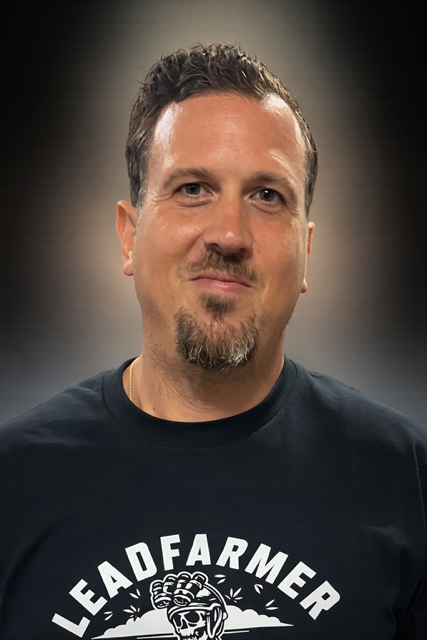 A man with short, dark hair and a goatee, wearing a black T-shirt with a slogan and graphic, standing in front of a plain background.