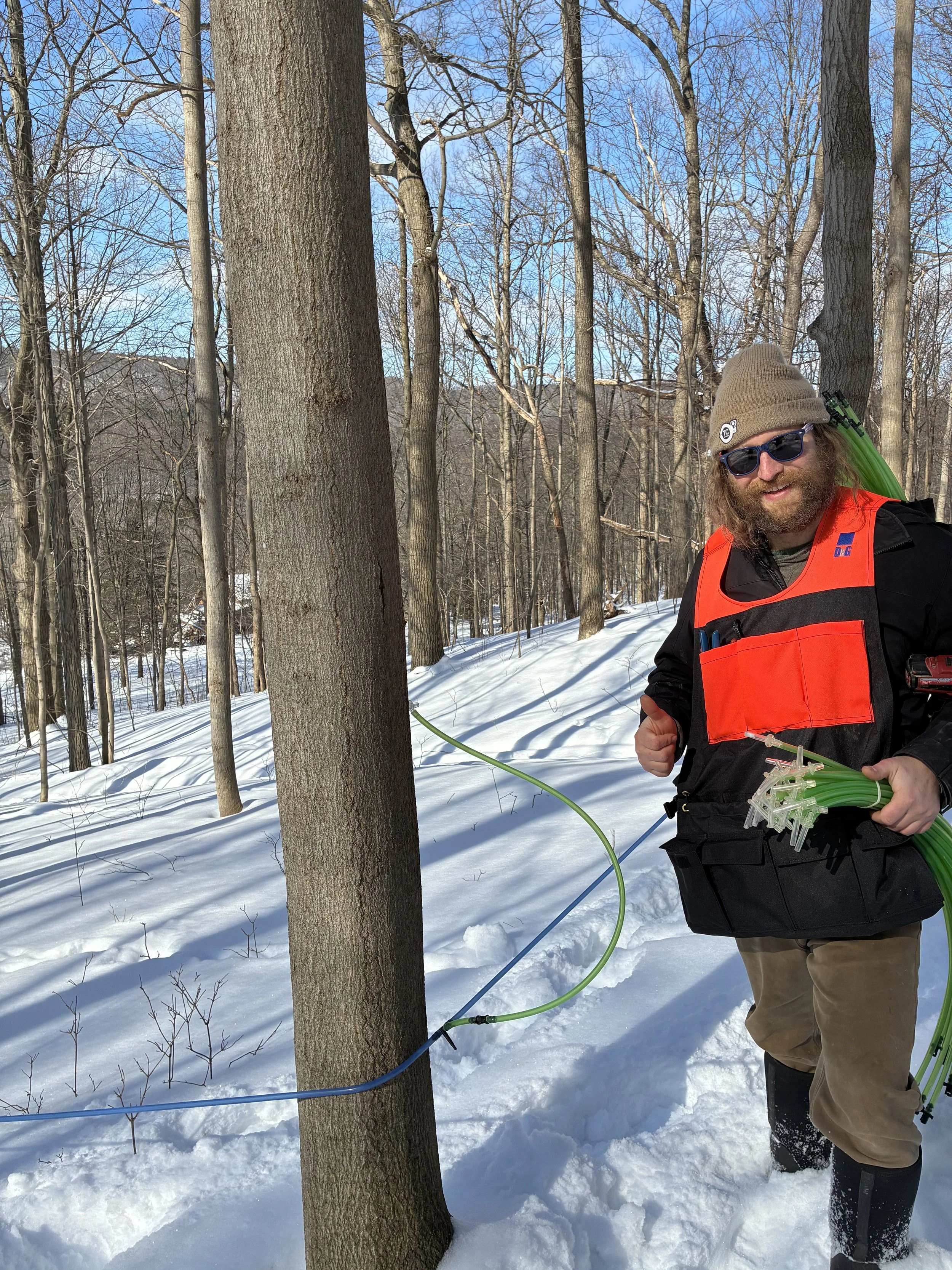 A man with long hair, beard, sunglasses, and a gray beanie hat standing in a snowy forest, holding green tubing and equipment, wearing a black jacket with a red vest and tan pants.