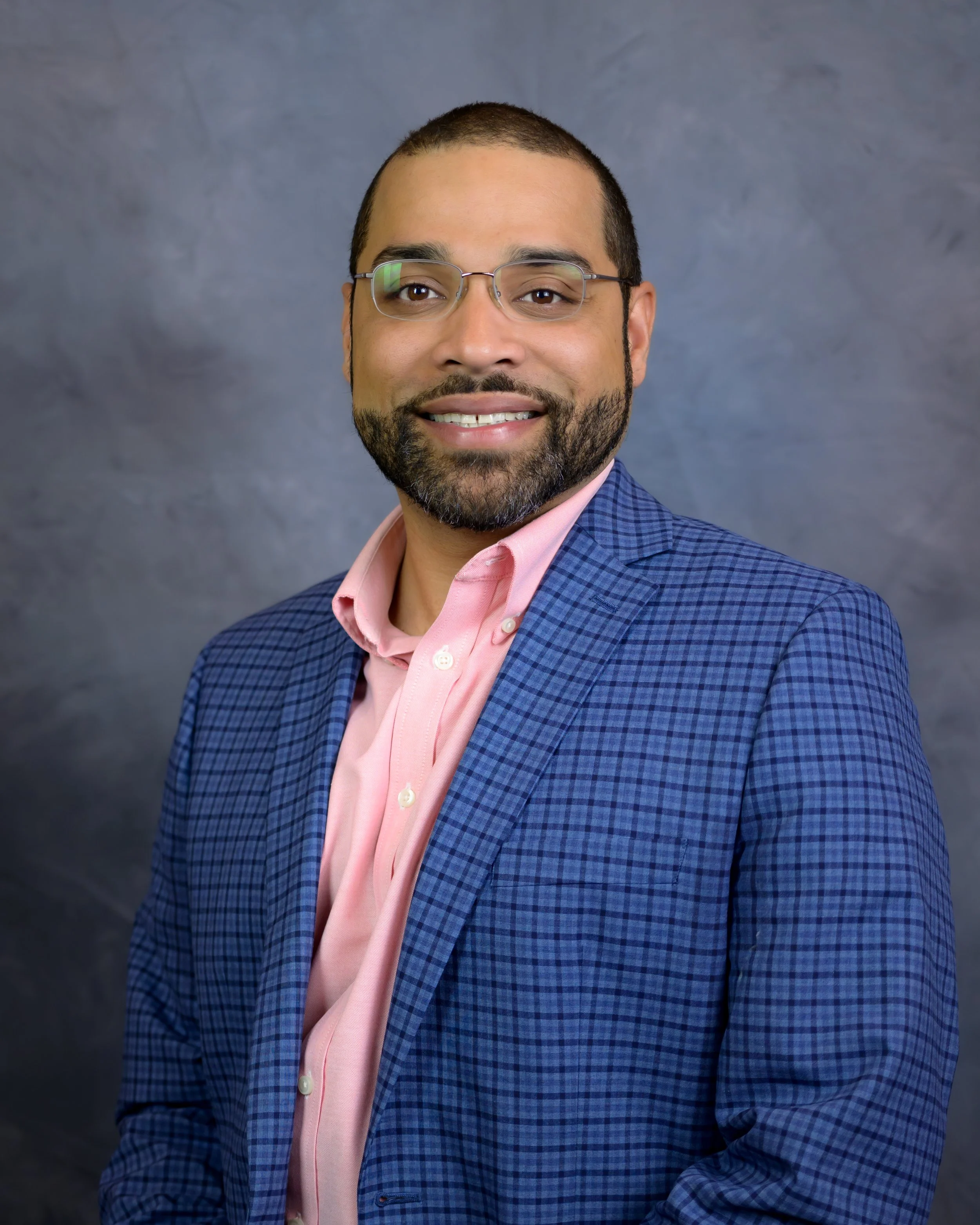 A man with glasses and a beard wearing a pink shirt and blue checkered blazer, smiling in front of a dark gray background.