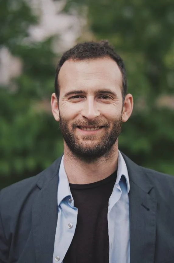A man with a beard and short dark hair, smiling, wearing a dark blazer over a light blue shirt and a black T-shirt, outdoors with a blurred green background.