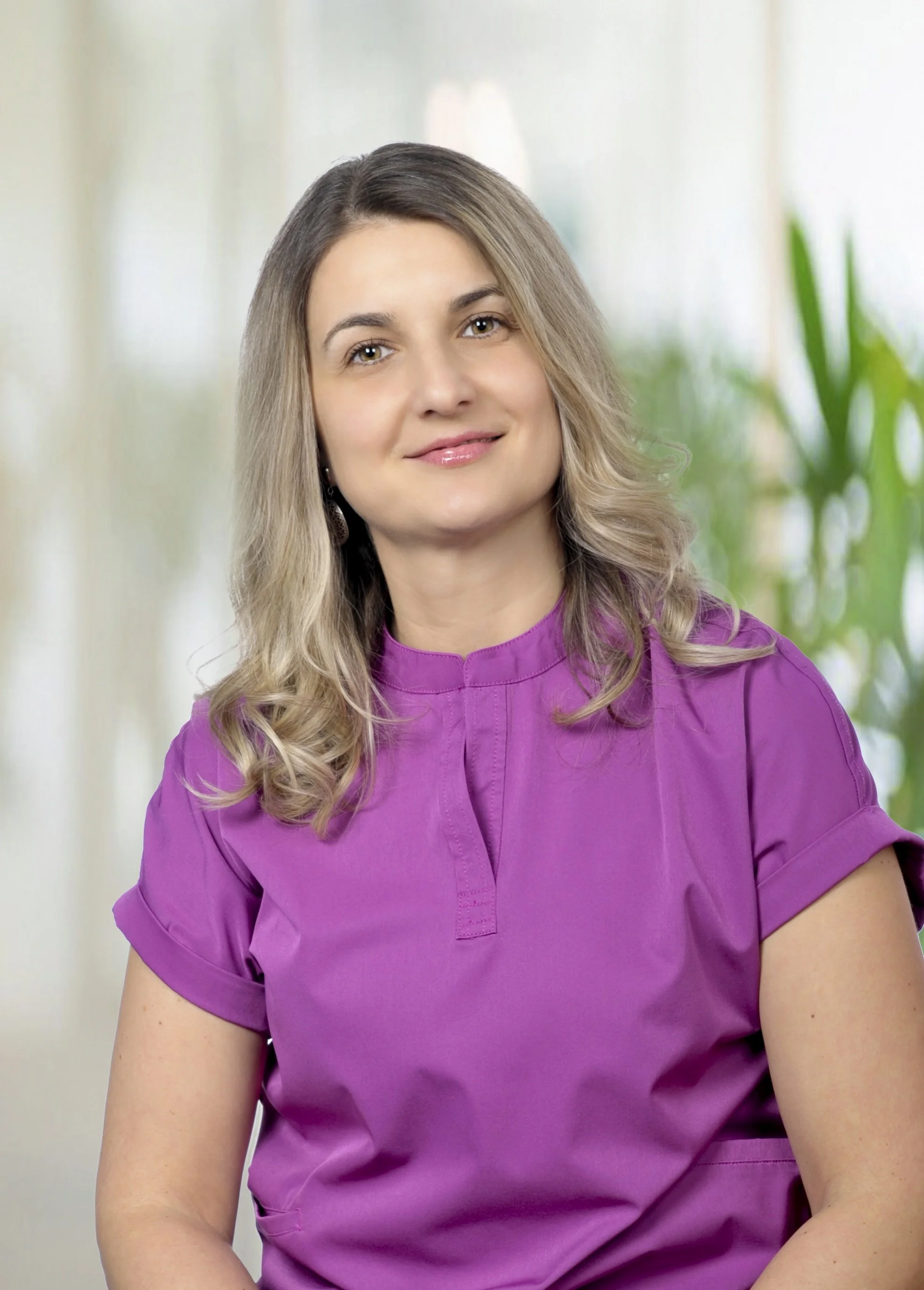 A woman with shoulder-length wavy blonde hair, wearing a purple shirt, smiling softly, sitting indoors with a blurred background of natural light and greenery.