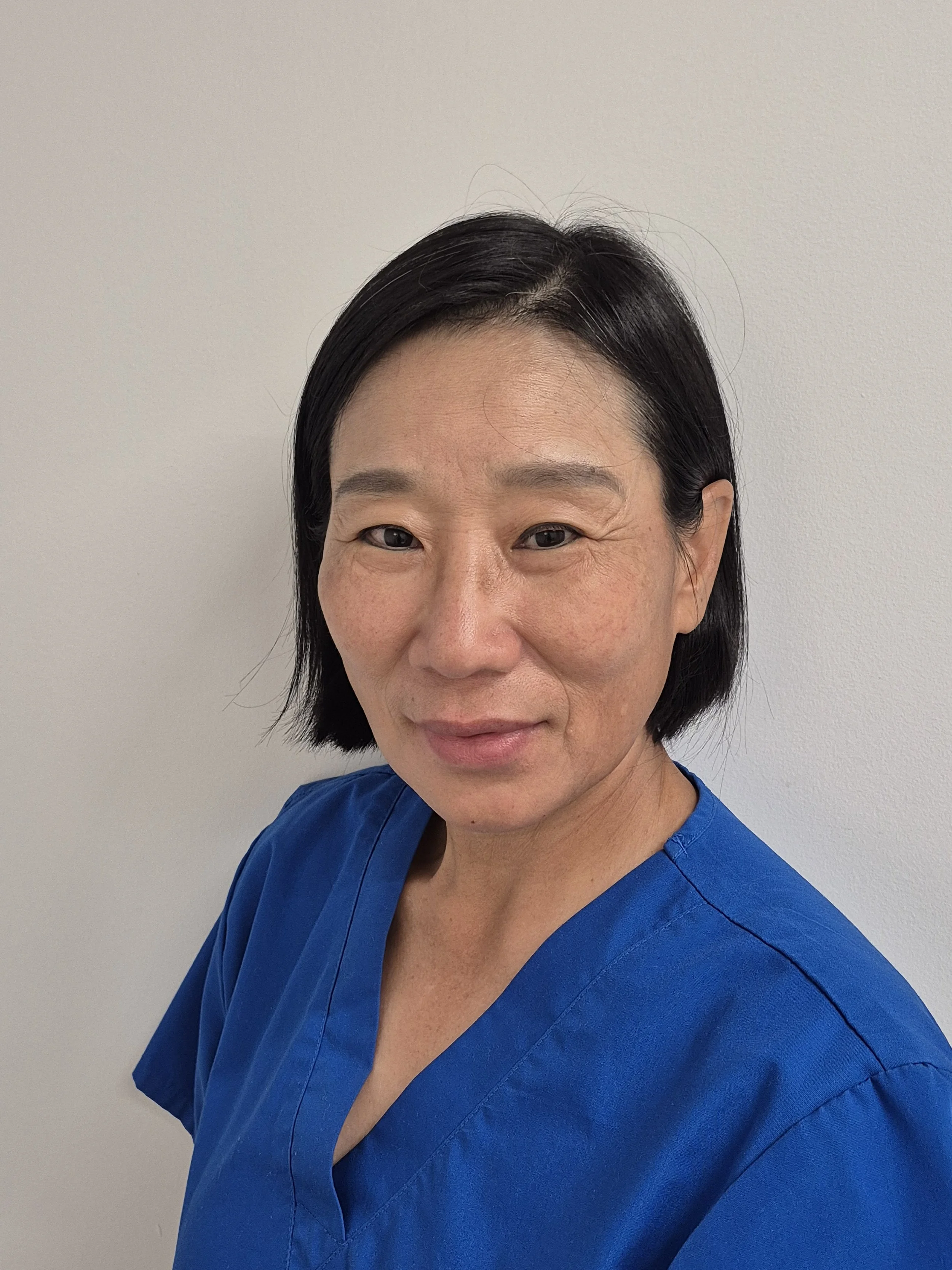 A woman with short black hair wearing blue scrubs standing in front of a plain white wall.