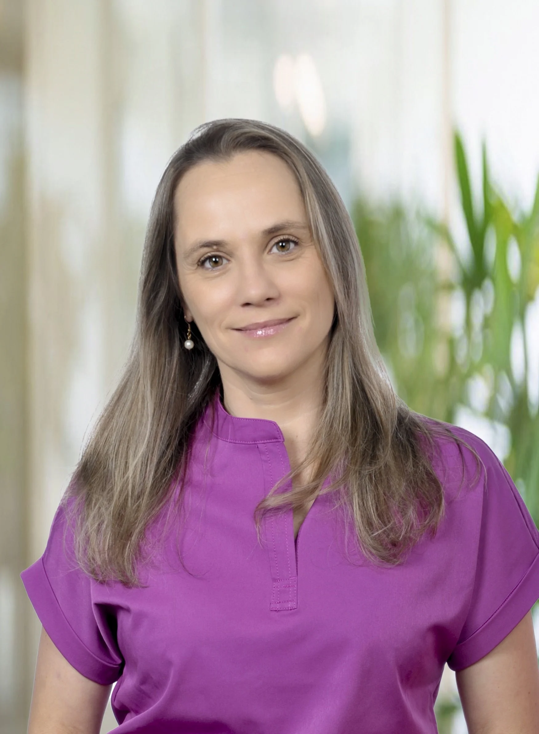 A woman with long brown hair wearing a purple shirt and pearl earrings, smiling in an indoor setting with green plants in the background.