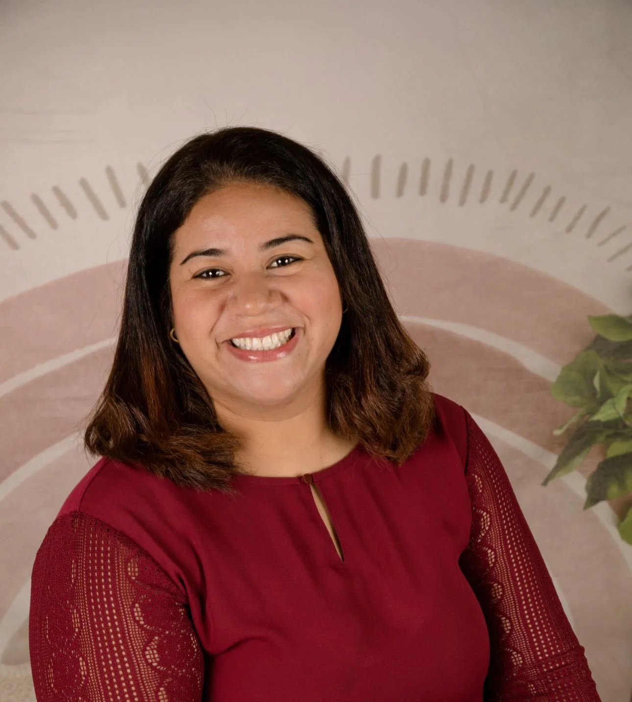 A woman with shoulder-length brown hair, wearing a red top with lace sleeves, smiling in front of a neutral background with a partial plant visible on the right.