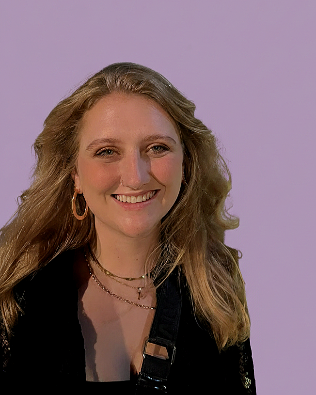 A woman with wavy, blonde hair, smiling, wearing hoop earrings, layered necklaces, and a black outfit, standing against a purple background.