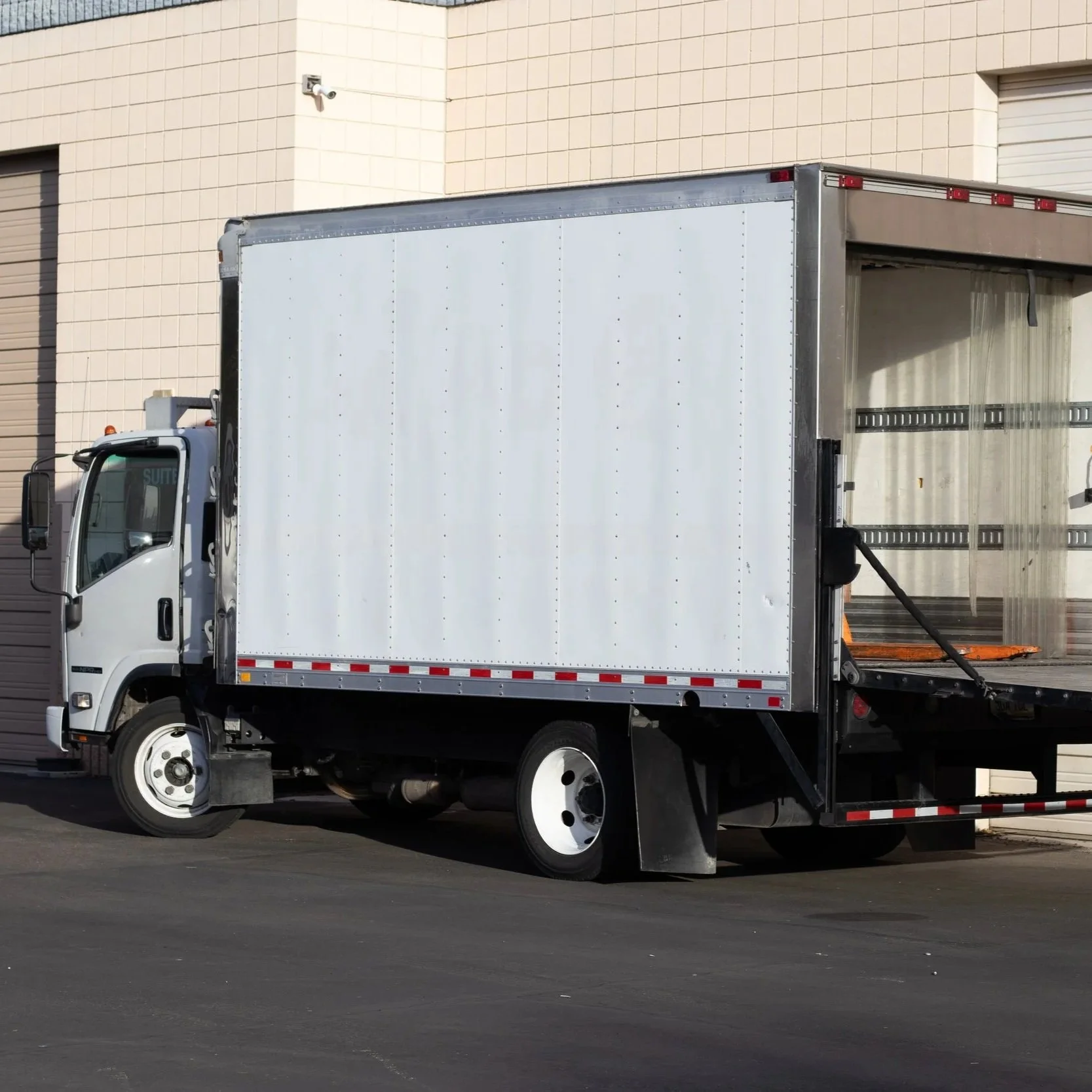 A white box truck with a blank cargo area parked on a street in front of a beige building with garage doors.