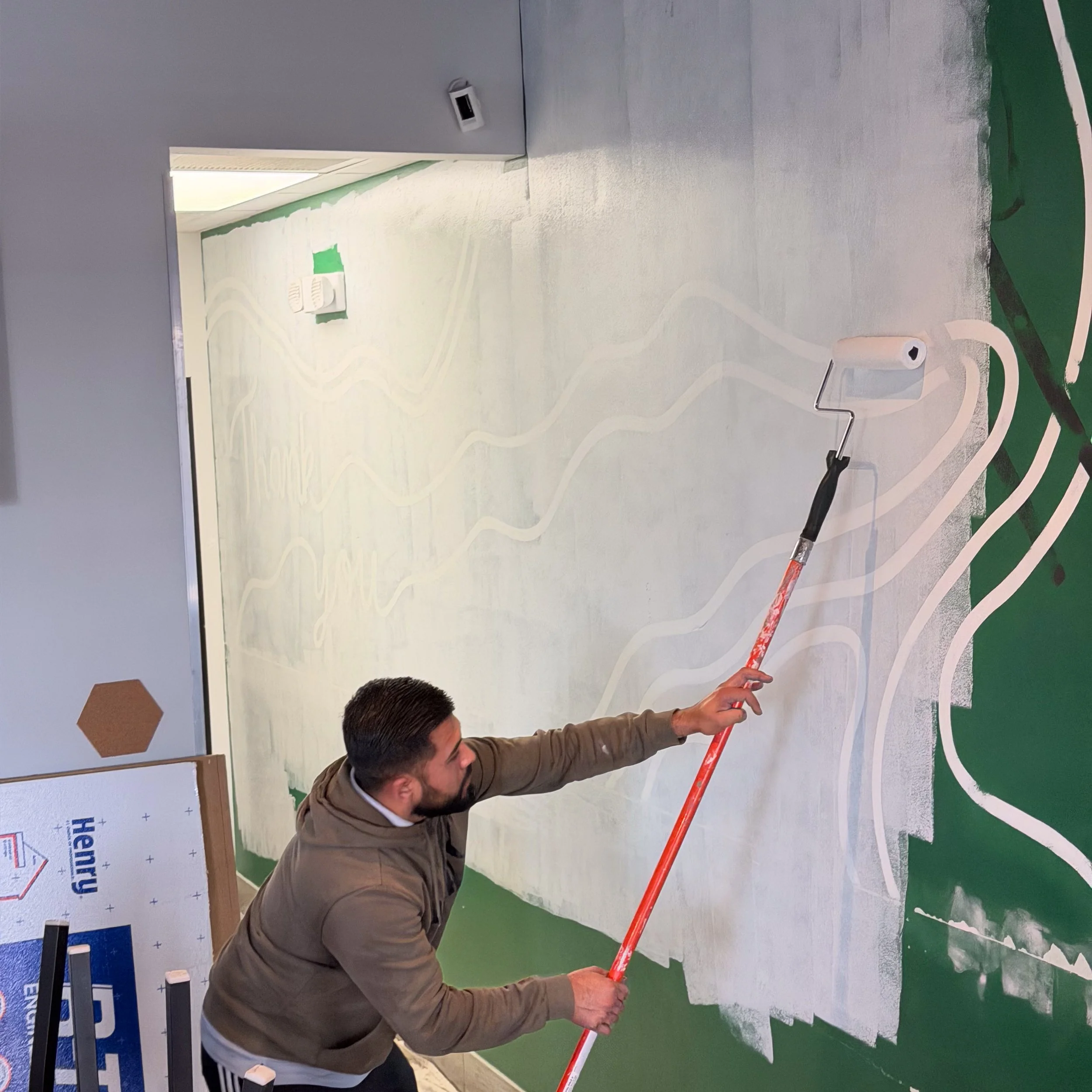 A man painting a mural on a wall with a paint roller on an extension pole. The wall has a design with wavy white lines.