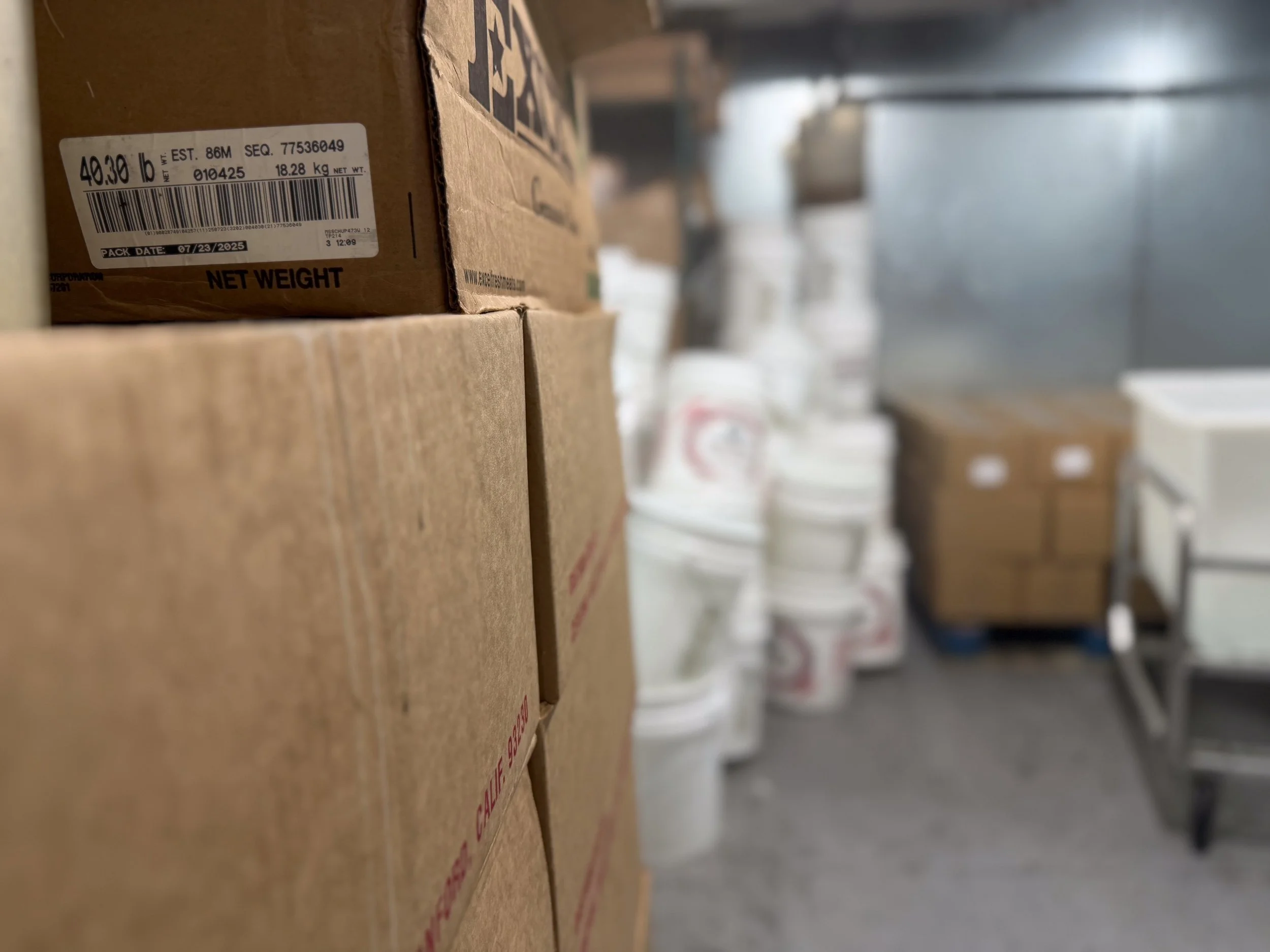 Close-up of stacked cardboard boxes with a label showing weight and barcodes, in a storage area with white buckets and other boxes in the background.