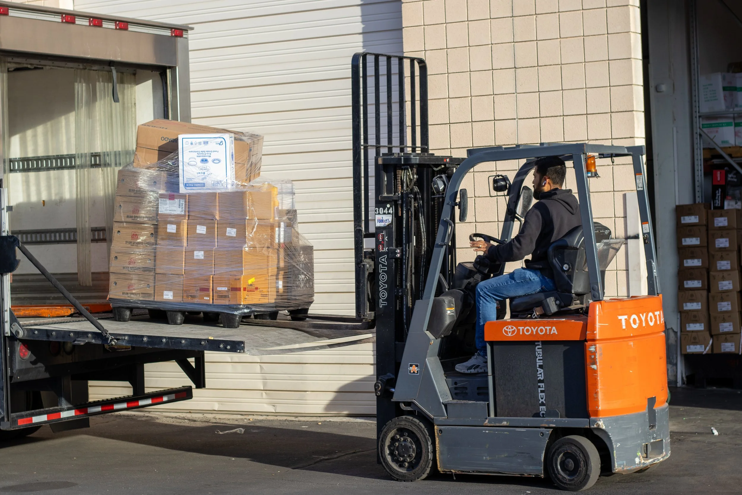 A man operating a Toyota forklift loading a pallet of boxes into a trailer outside a warehouse.