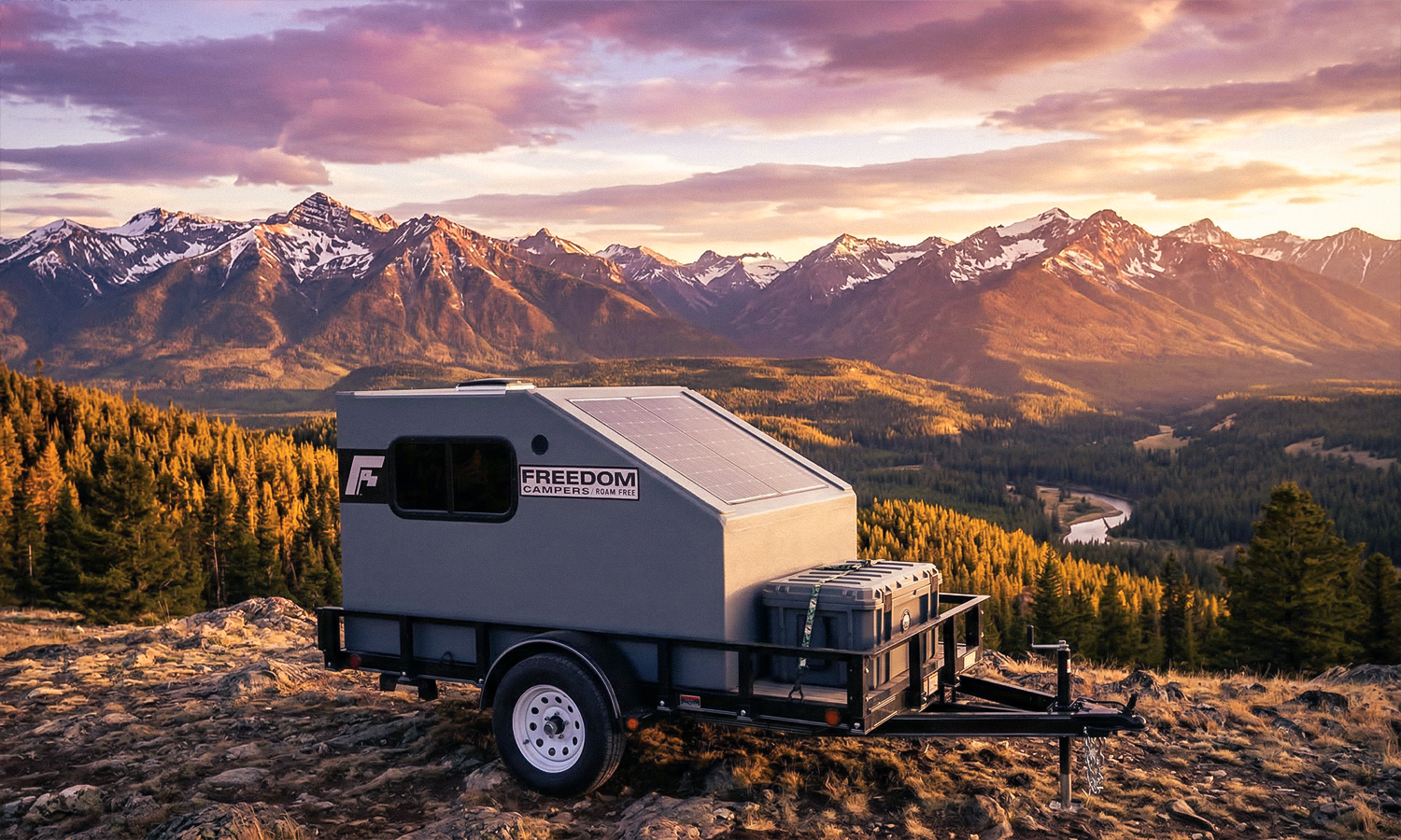 A camper trailer with solar panels set up on a rocky hilltop overlooking a sunset mountain landscape with forested valleys and a winding river.