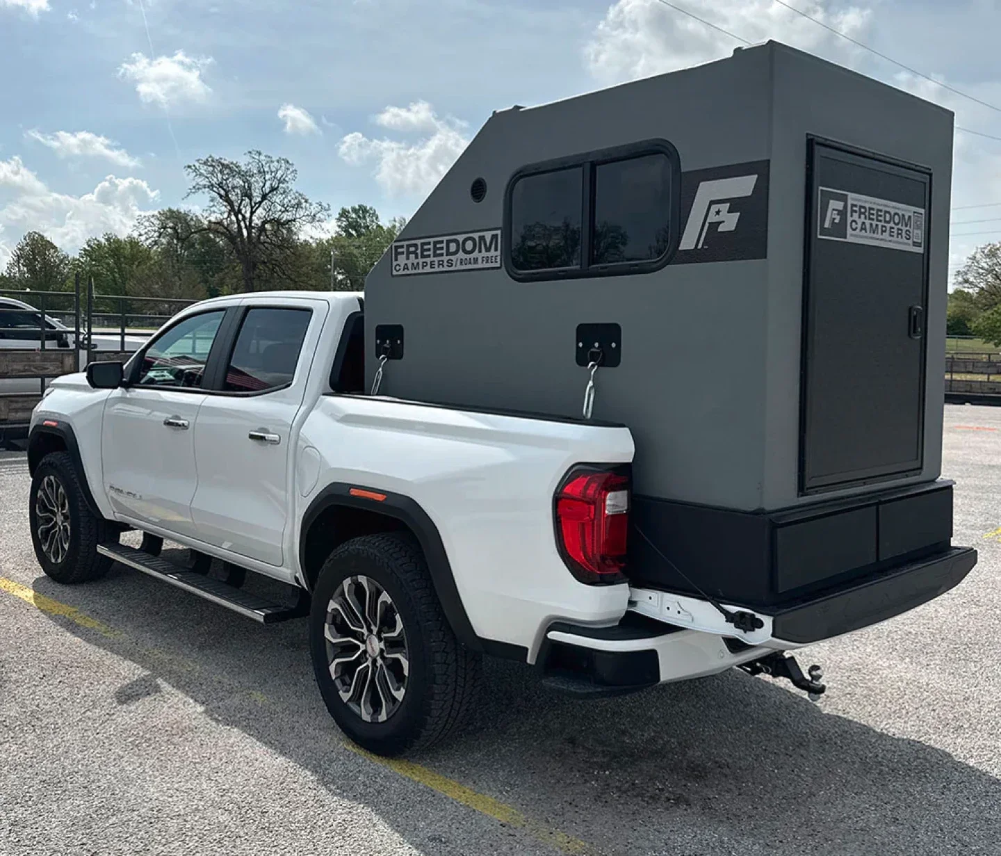 White pickup truck with a camper shell attached to the bed, parked in a lot under a partly cloudy sky.