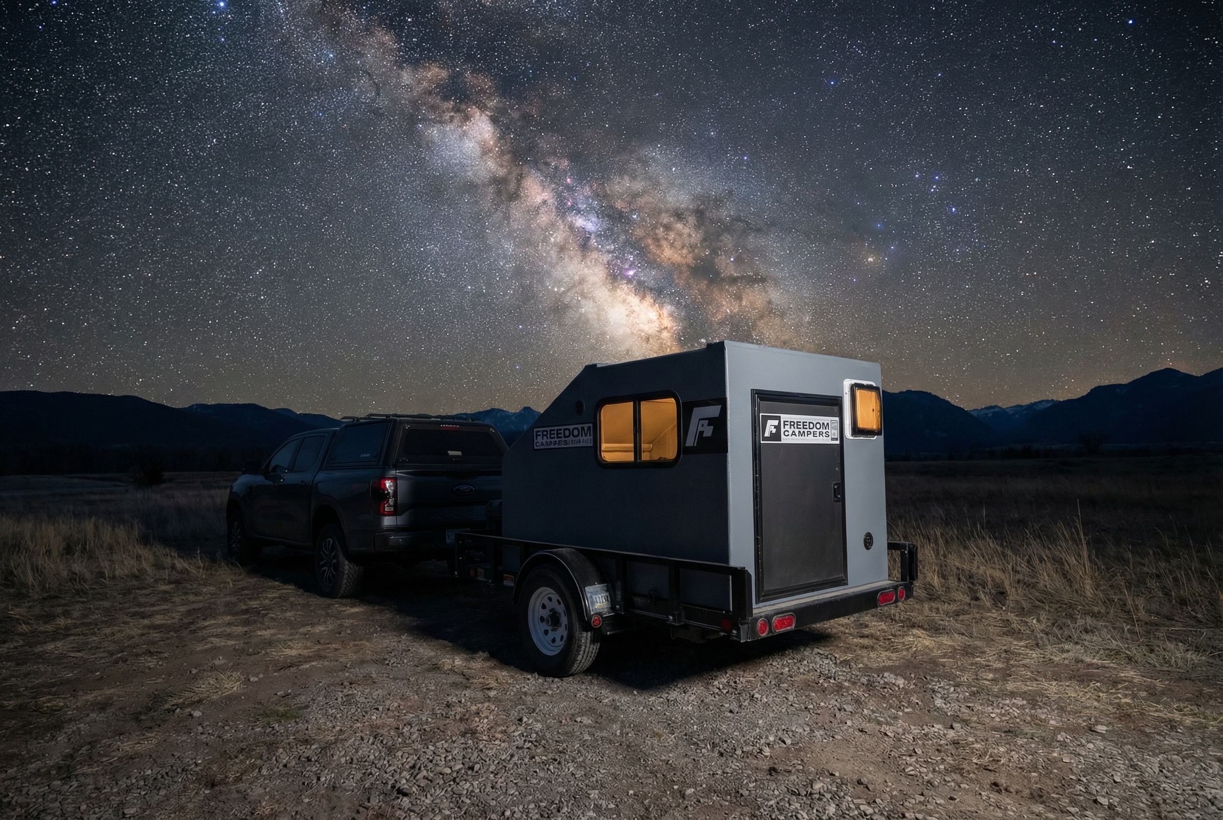 A black pickup truck towing a small, gray camping trailer with illuminated interior, parked on a dirt field under a clear night sky filled with stars and the Milky Way galaxy.
