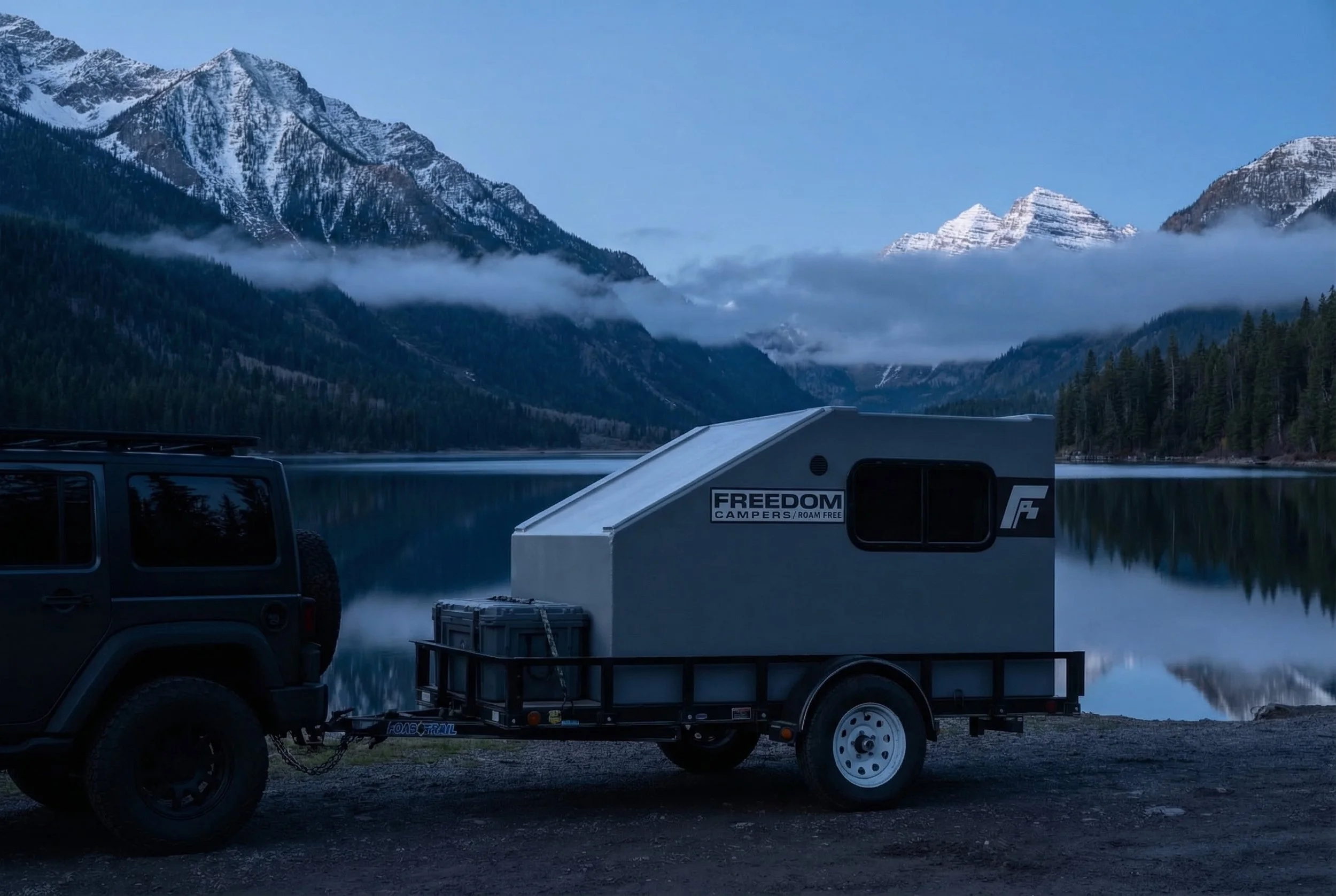 A black SUV towing a small camper trailer parked near a serene mountain lake with snow-capped mountains and clouds in the background