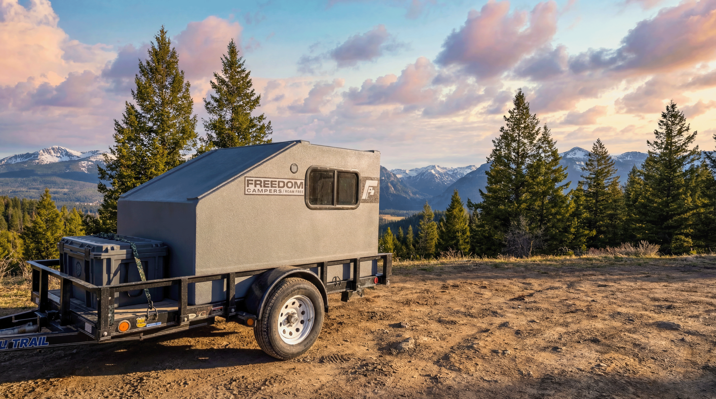 A small camper trailer parked on a dirt patch in a forested mountain landscape at sunset.