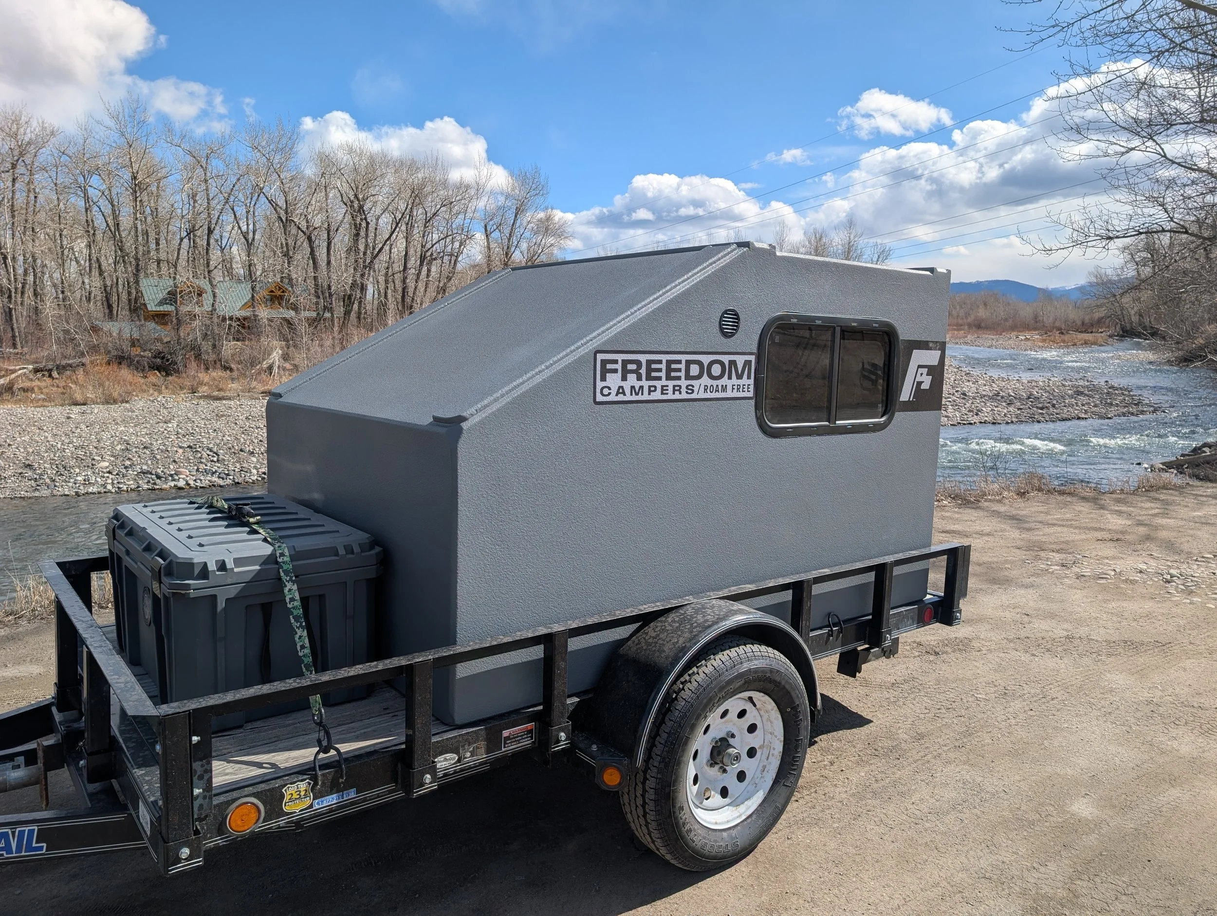 A small camper trailer with a gray exterior and a window, labeled 'FREEDOM CAMPERS / ROAM FREE', parked on a dirt area near a river with leafless trees and a blue sky with clouds in the background.