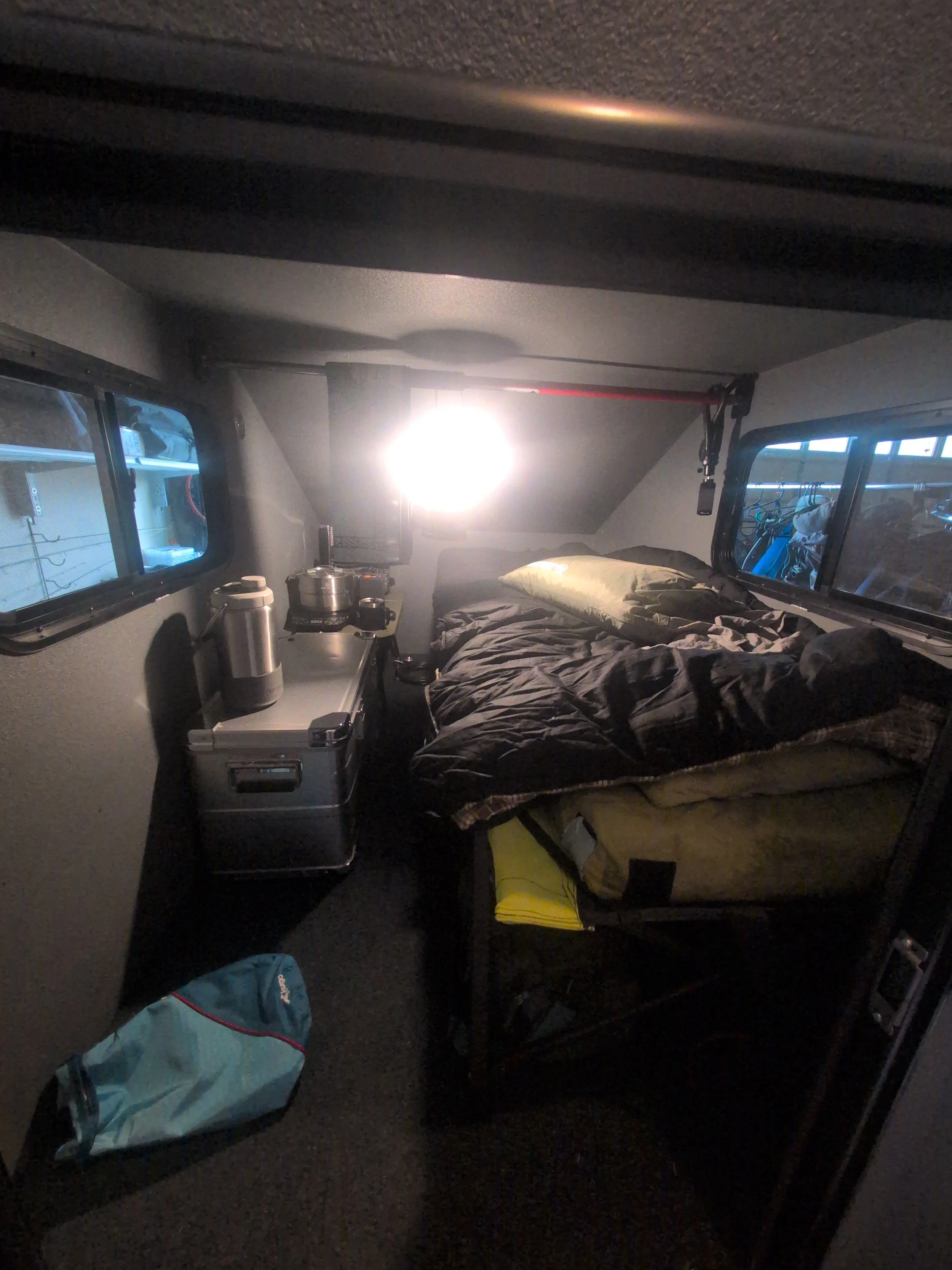 Inside a compact camper van or RV with a small bed, illuminated by a bright light at the head of the bed. There are windows on either side, a small shelf or counter with a kettle and containers, and a bag on the floor.